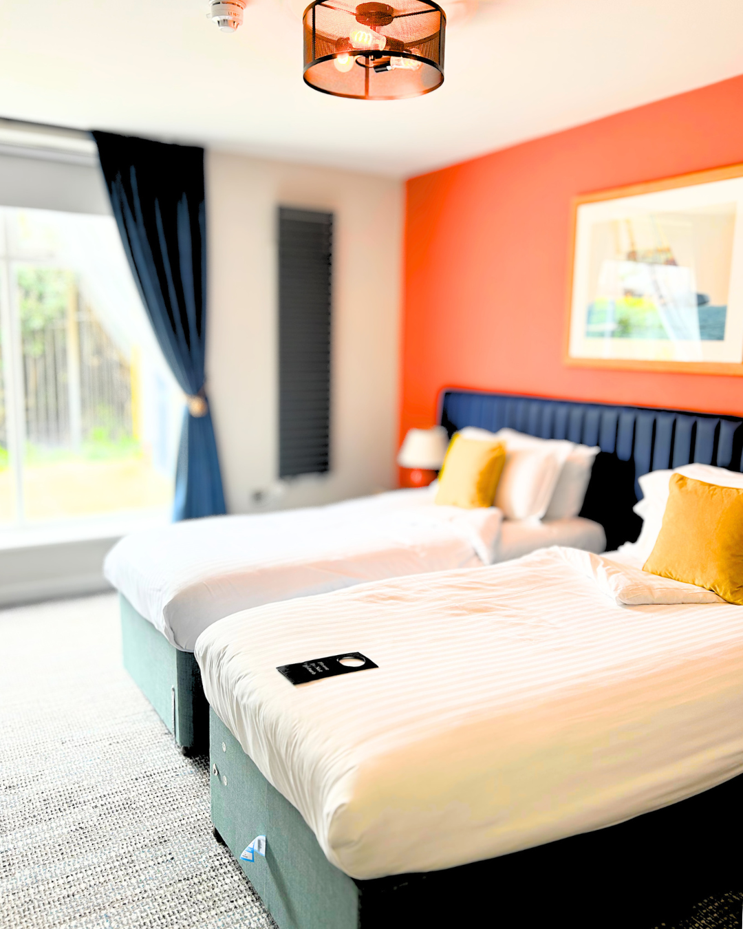 Two single beds with white bedding and yellow pillows in a bedroom, with a large window with dark blue curtains and a framed artwork on an orange accent wall.