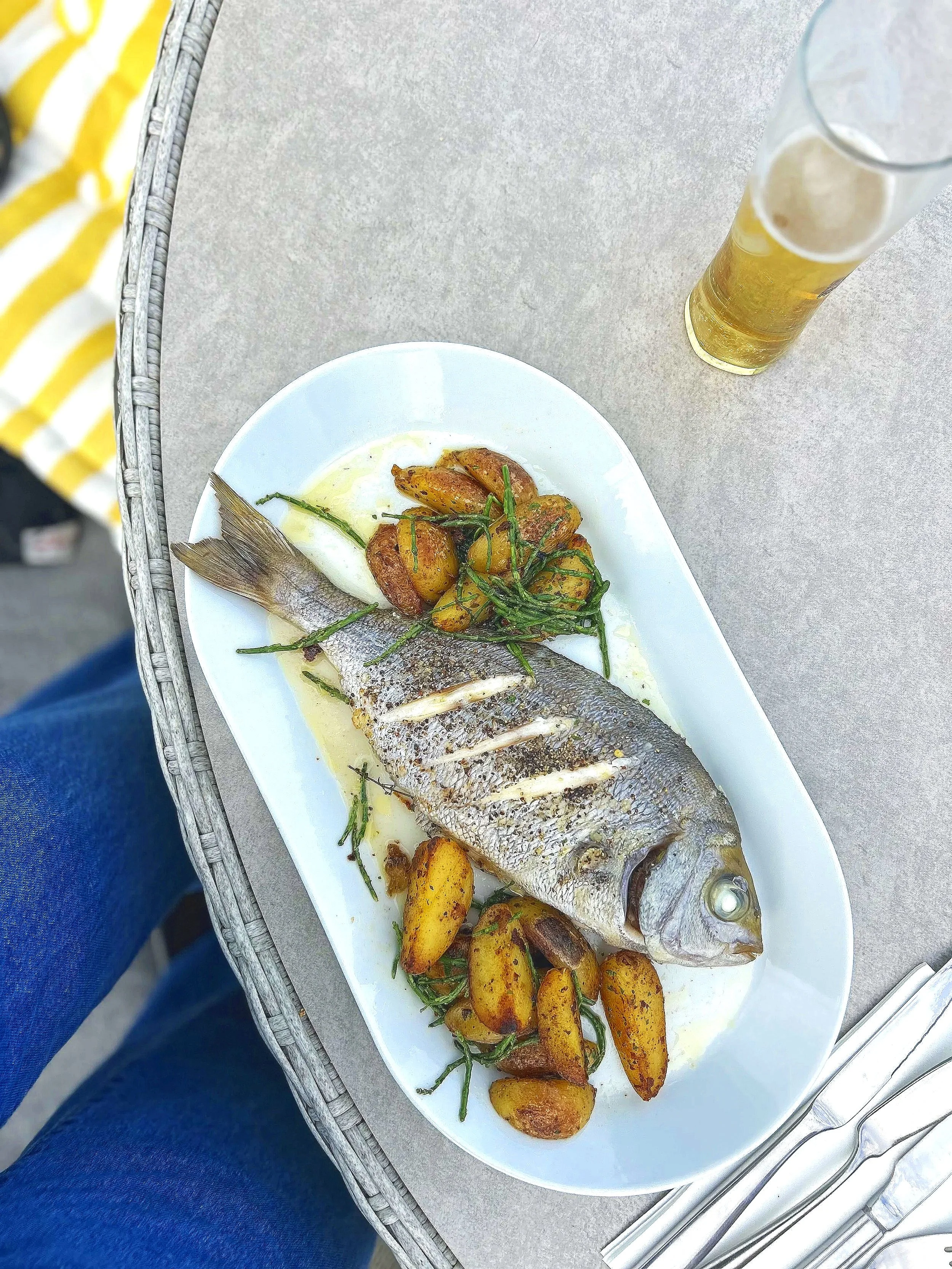 A plate with a whole cooked fish, topped with green herbs, accompanied by roasted potatoes and green beans, served with a side of yellow sauce. There is a glass of beer on the table next to the plate.