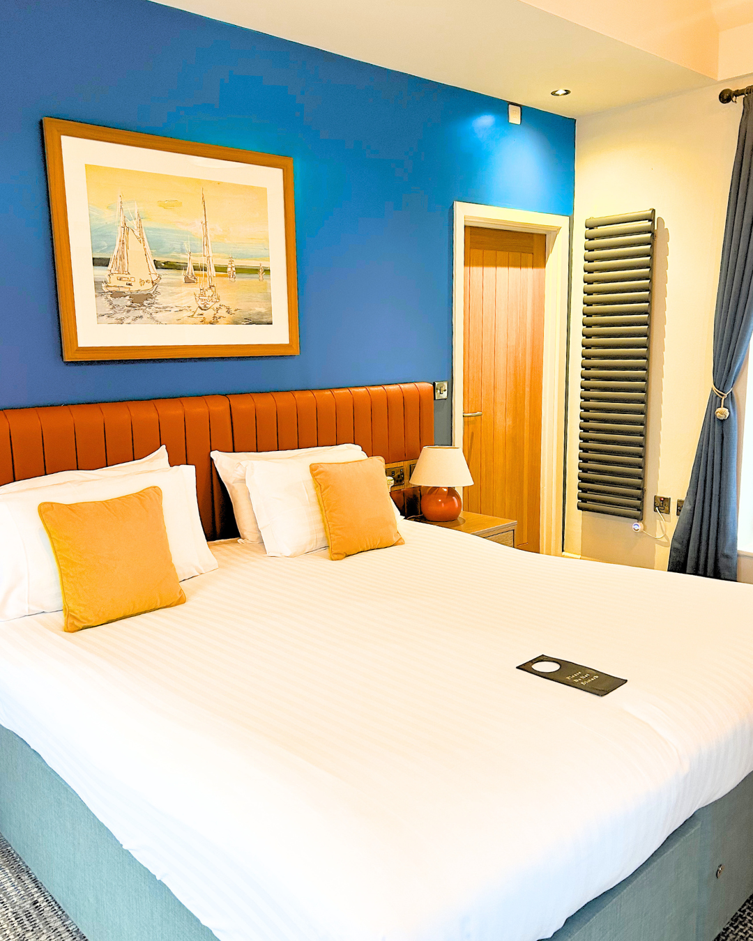 A neatly made bed with white sheets and three pillows, two with orange pillowcases and one with a white pillowcase, in a hotel room. There's a wooden headboard, a small bedside table with a lamp, a blue wall with a framed painting of sailboats, and a wooden door in the background. To the right, there's a window with a curtain and a modern radiator.