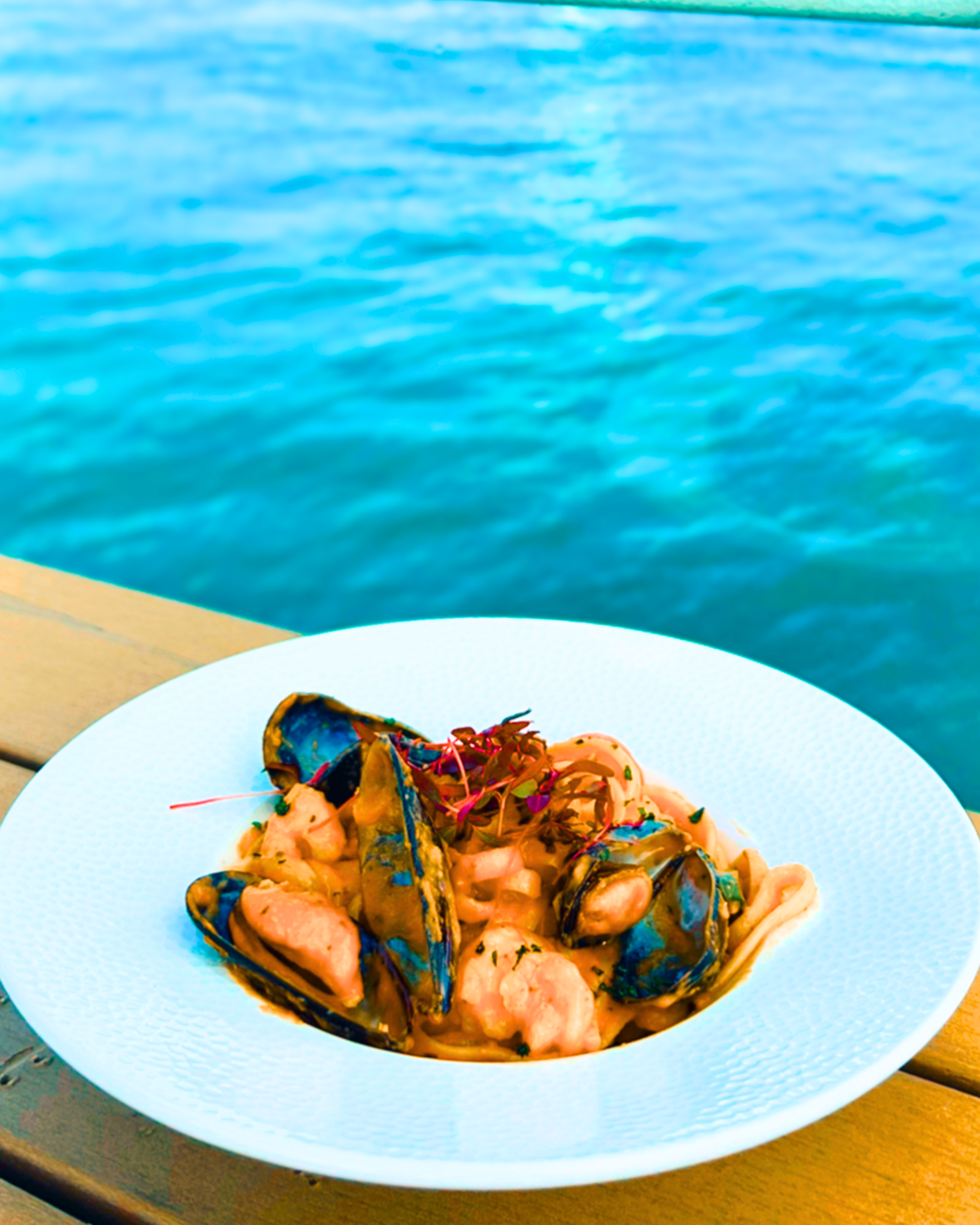 Seafood pasta dish with mussels, shrimp, and herbs served on a white plate, with a body of water in the background.