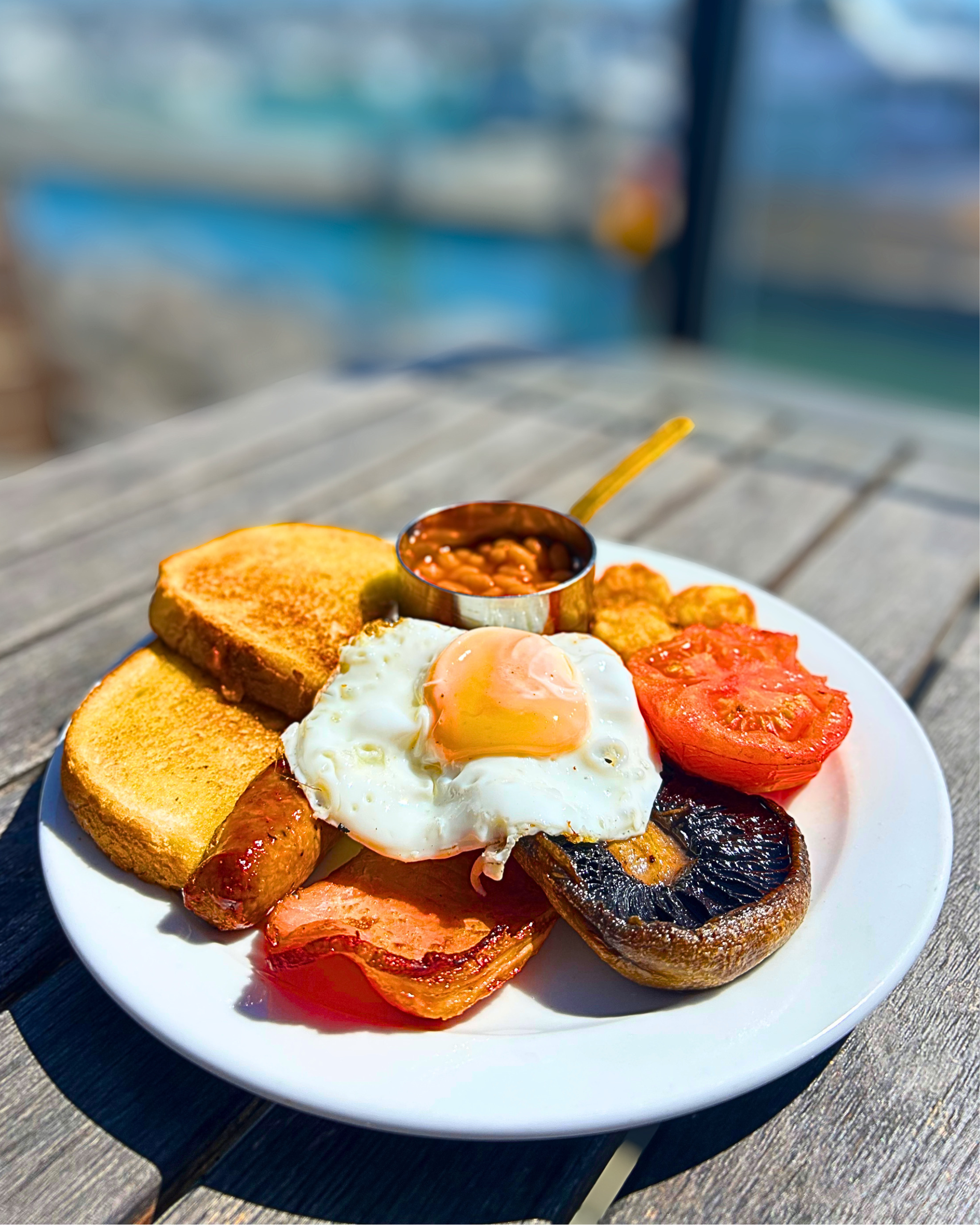 Plate of breakfast food including toast, baked beans, fried egg, sausage, tomato, mushroom, and bacon on a white plate.