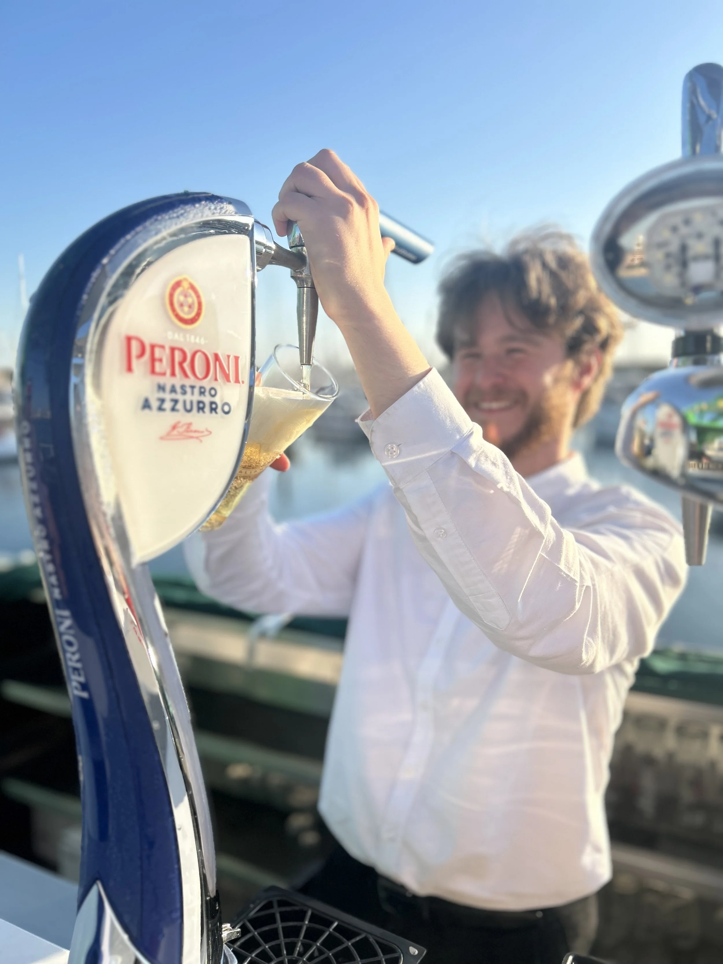 A man with curly hair and a white shirt is pouring Peroni beer into a glass from a tap at an outdoor setting with a clear blue sky.