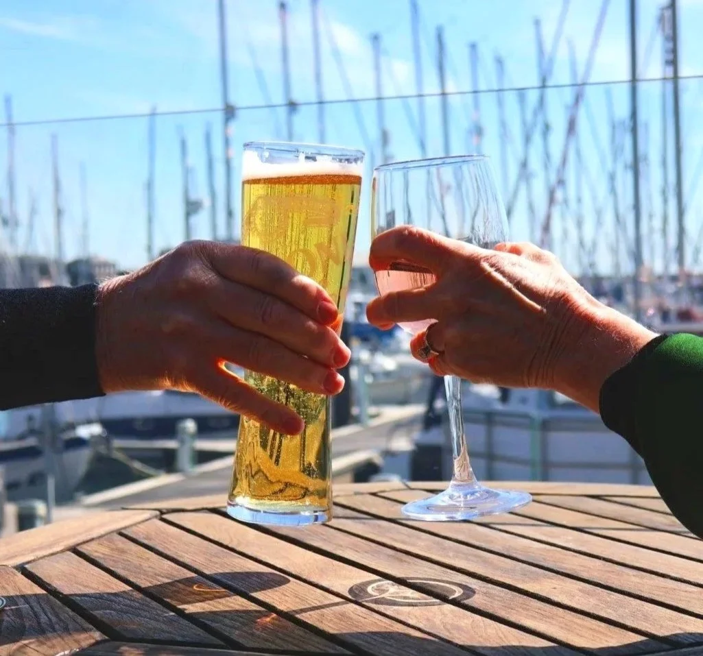 Two people clinking glasses, one with beer and the other with wine, at an outdoor marina with boats and masts in the background.