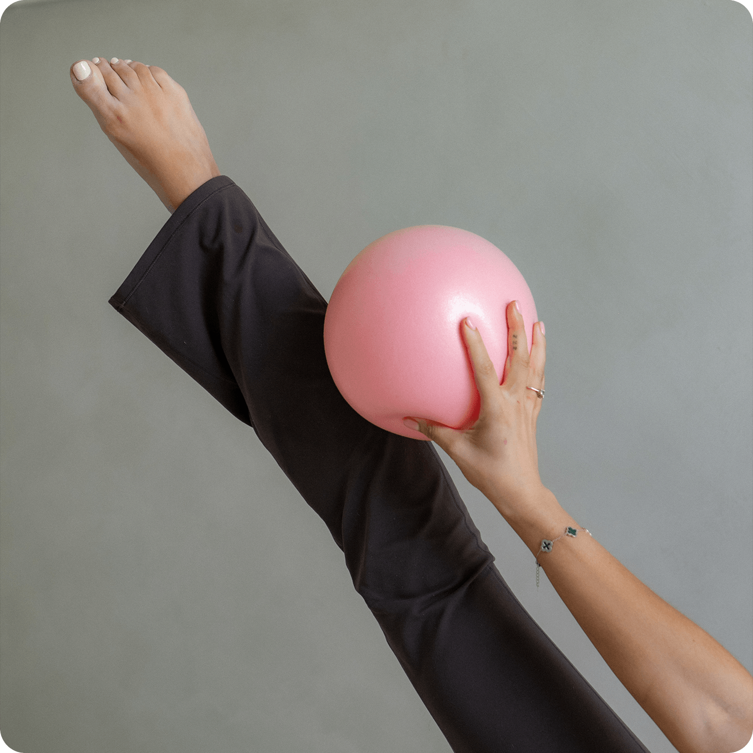 A person holding a pink exercise ball against their leg, which is extended upward, with a gray wall in the background.