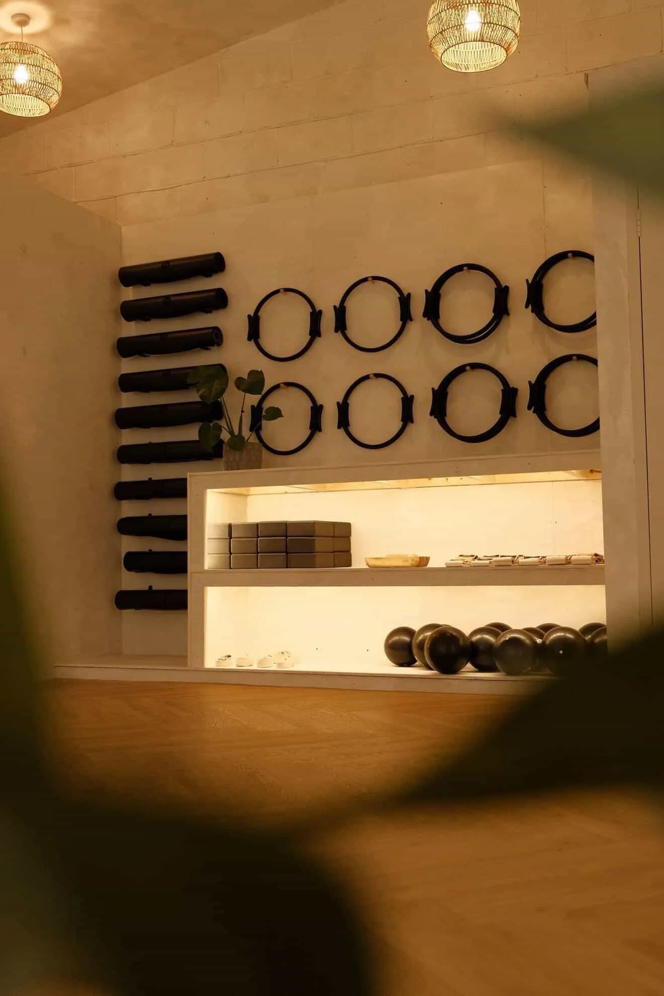 Interior space with wooden walls and ceiling, featuring black cylindrical objects on the left and black circular bands hanging on the wall. There is a white illuminated shelf with stacked items and a small plant at the corner. Large black balls are arranged at the bottom of the image.