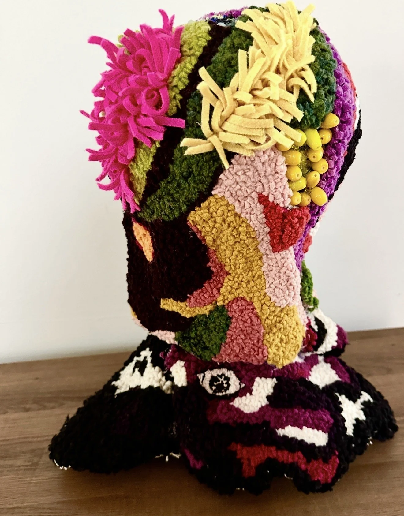 Abstract rug hooking bust sculpture representing a person, decorated with various yarns, ribbons, and beads, placed on a wooden surface against a white background.