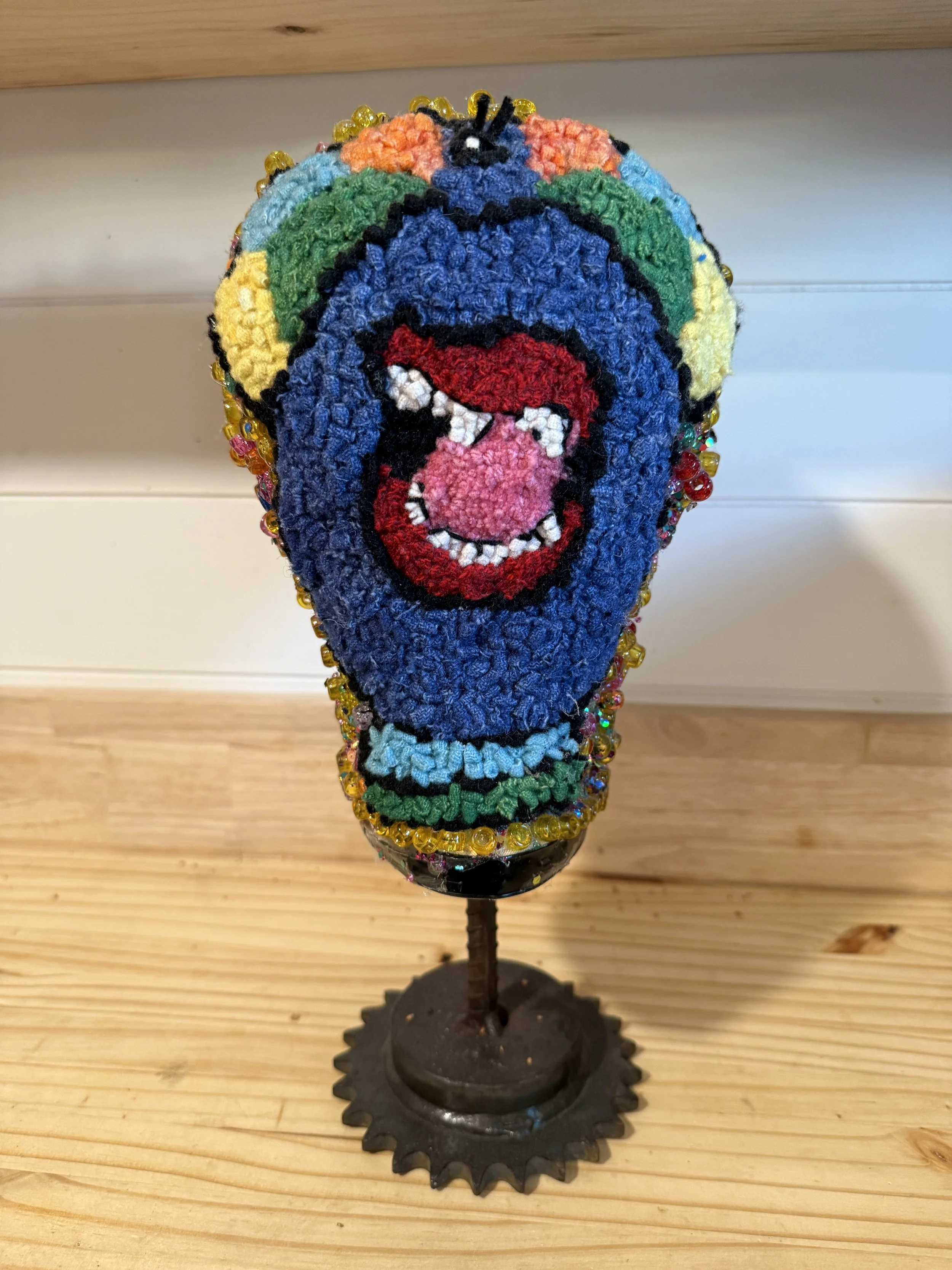 Colorful sculpture/ art piece featuring a smiling face with a pink nose, red lips, and white teeth, decorated with beads and set on a stand.