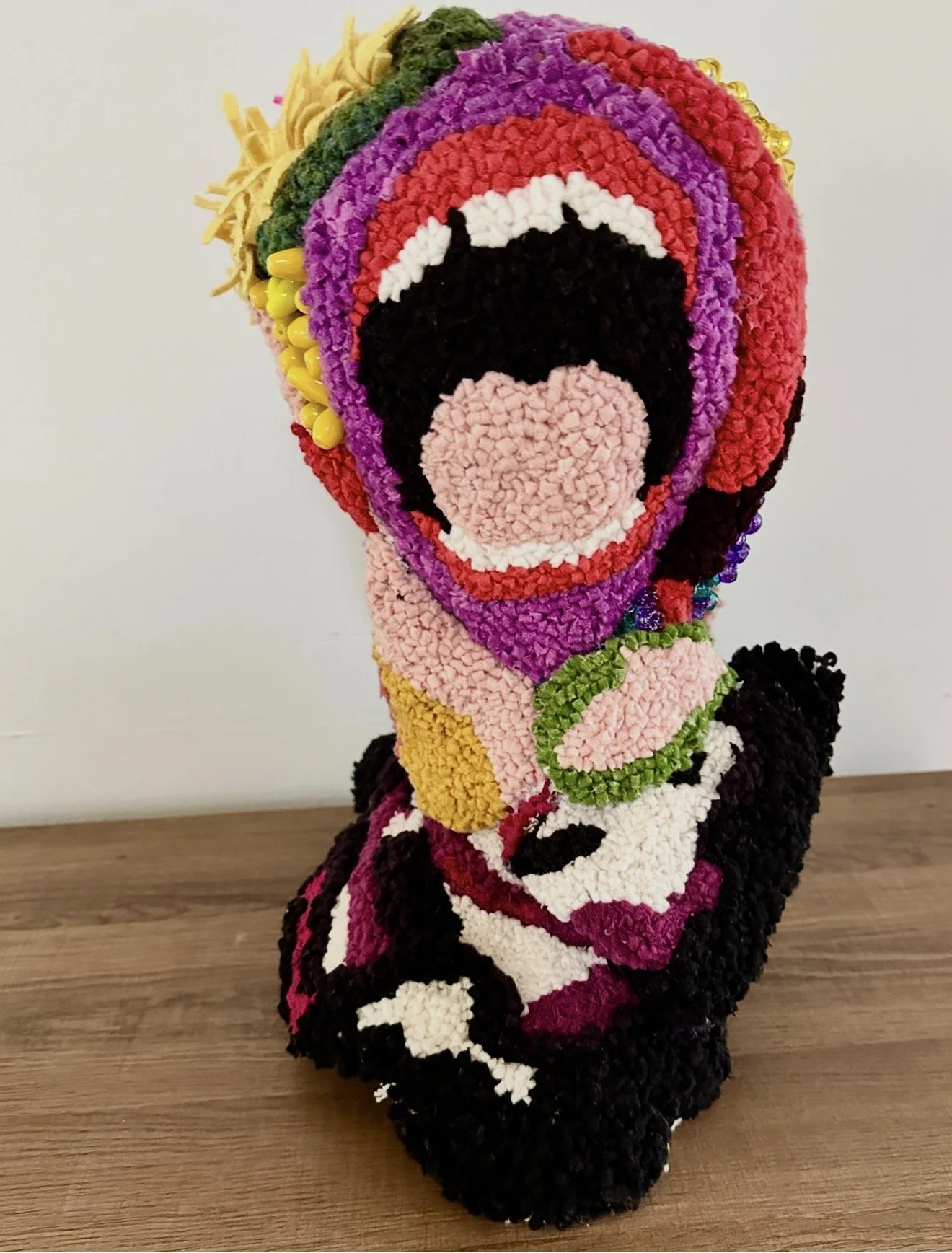 Colorful abstract portrait with pink hair, black eyes, green earrings, and a pink mouth, sitting on a wooden surface against a plain white background.