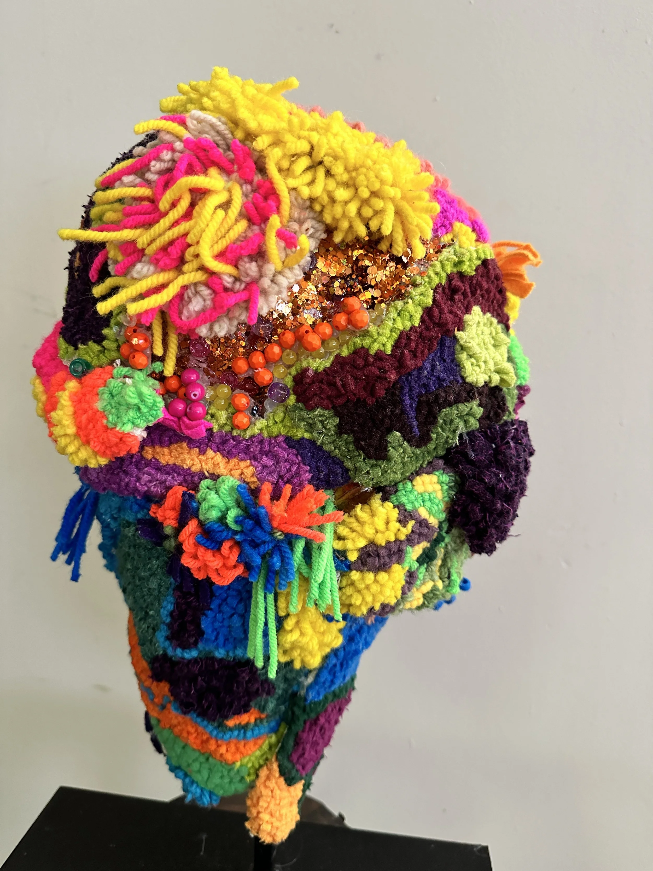 Colorful textile sculpture with yarn, beads, and sequins on a black stand against a plain wall.