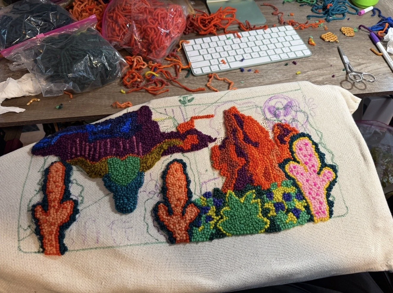A partially completed colorful  landscape with cacti, mountains, and desert scene, on a piece of fabric on a worktable surrounded by sewing and craft supplies.