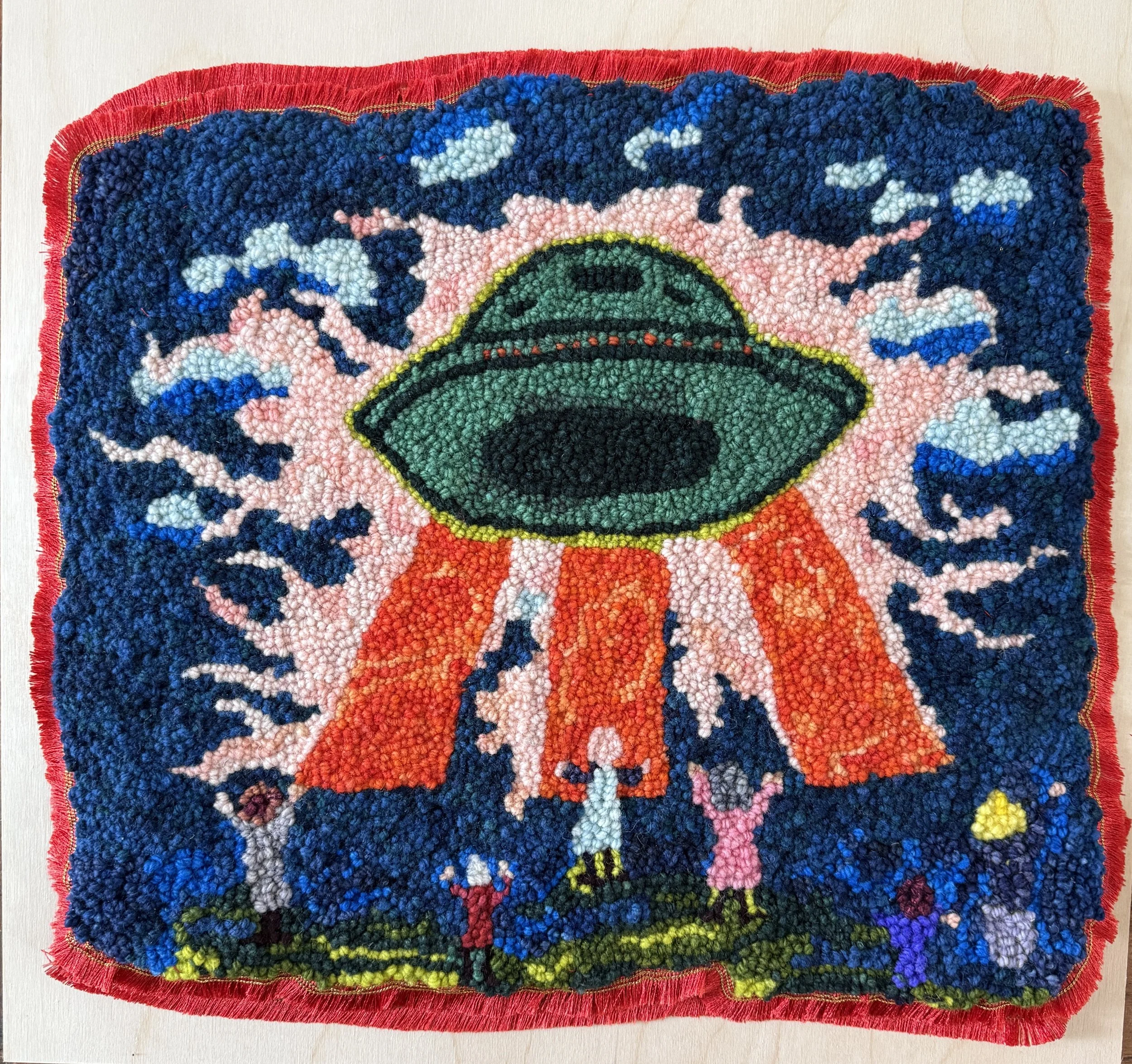 A close-up of a colorful, textured rug or artwork with a vibrant design featuring a green eye at the center, surrounded by pink, blue, and black patterns, with a fringed red border.