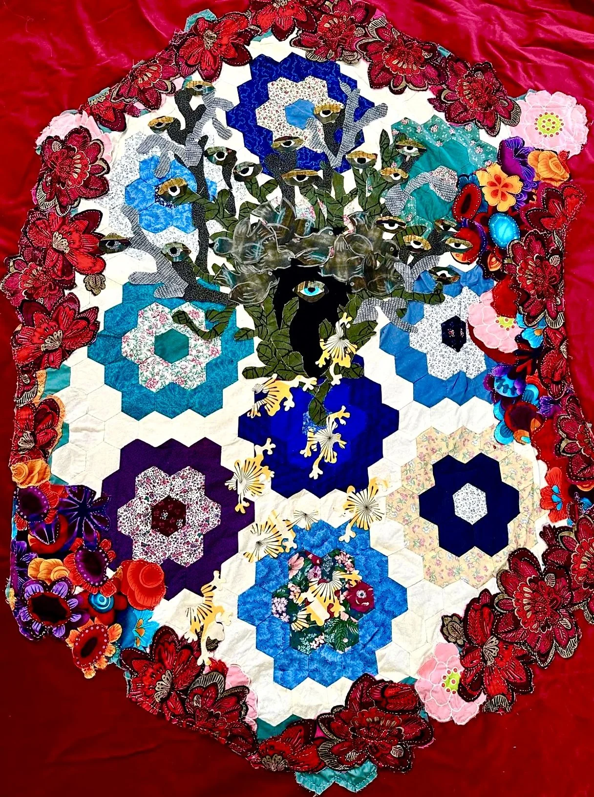 A quilt featuring a floral and geometric design with abstract  face motif, surrounded by hexagonal patterns and floral embellishments, with red fabric on the border.