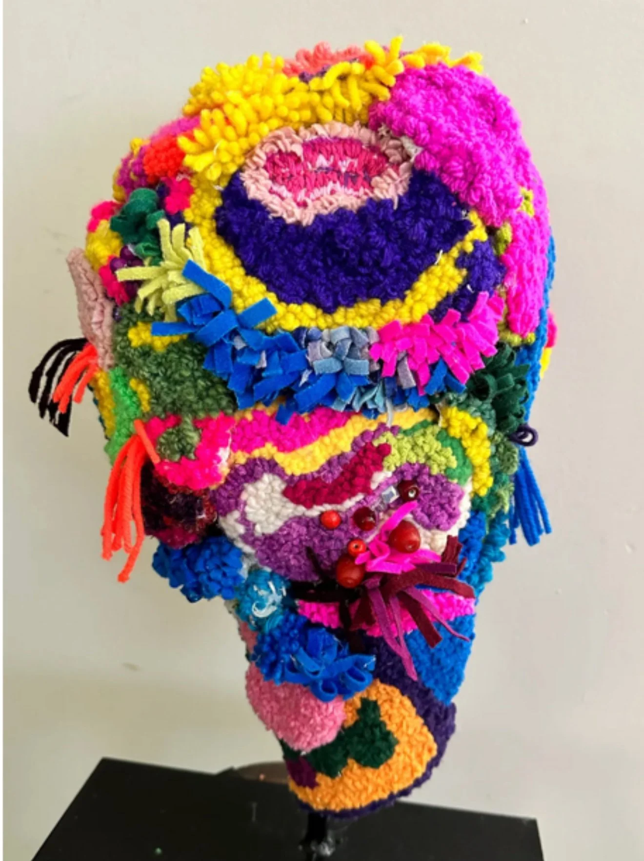 Colorful self portrait made of various yarn and fabric textures with flowers, bows, and decorative elements, standing on a black base against a plain background.