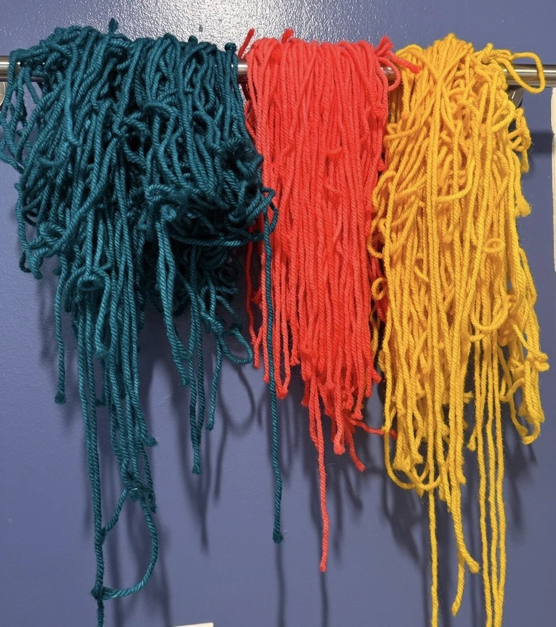 Three bunches of yarn in teal, red, and yellow hanging on a silver rod against a blue wall.