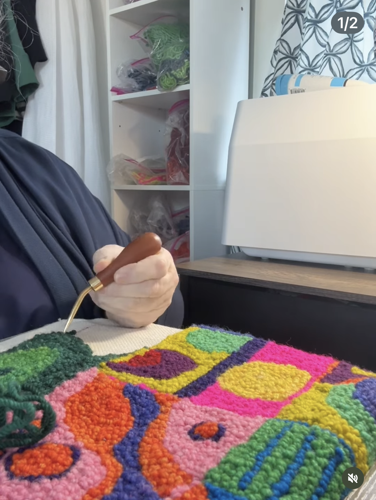 Person working on a colorful wool rug design sitting at a table. In the background, there is a white shelf with plastic bags of yarn or thread, white curtains, and a partly visible printer.
