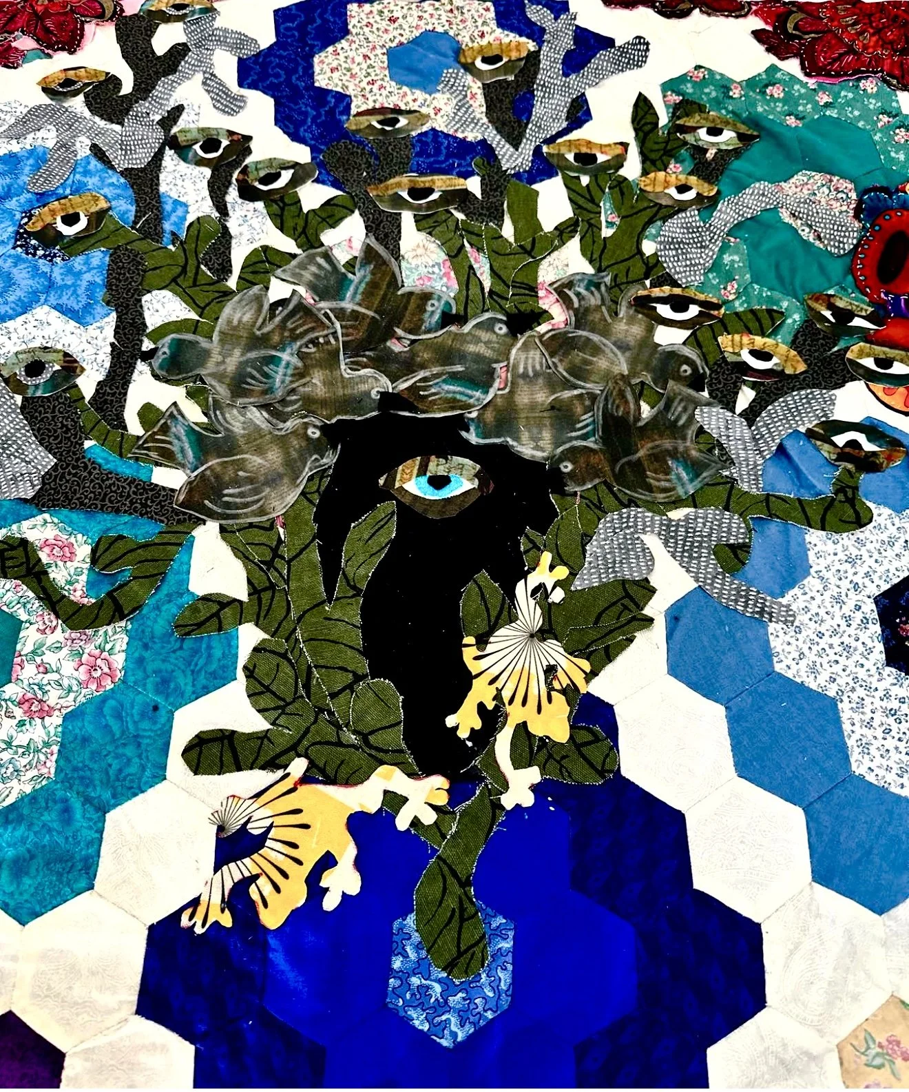 Fabric quilt displaying a stylized abstract face with flowers, leaves, birds, and earrings, constructed from various patterned fabric patches.