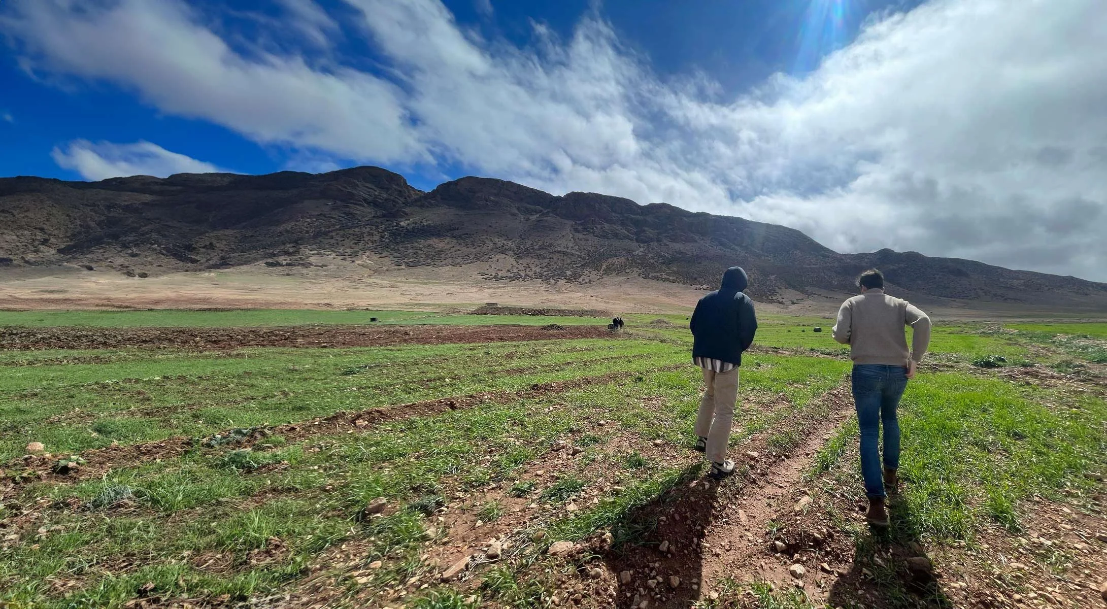 Two people walking on a dirt path through green fields with mountain range in background under partly cloudy sky.