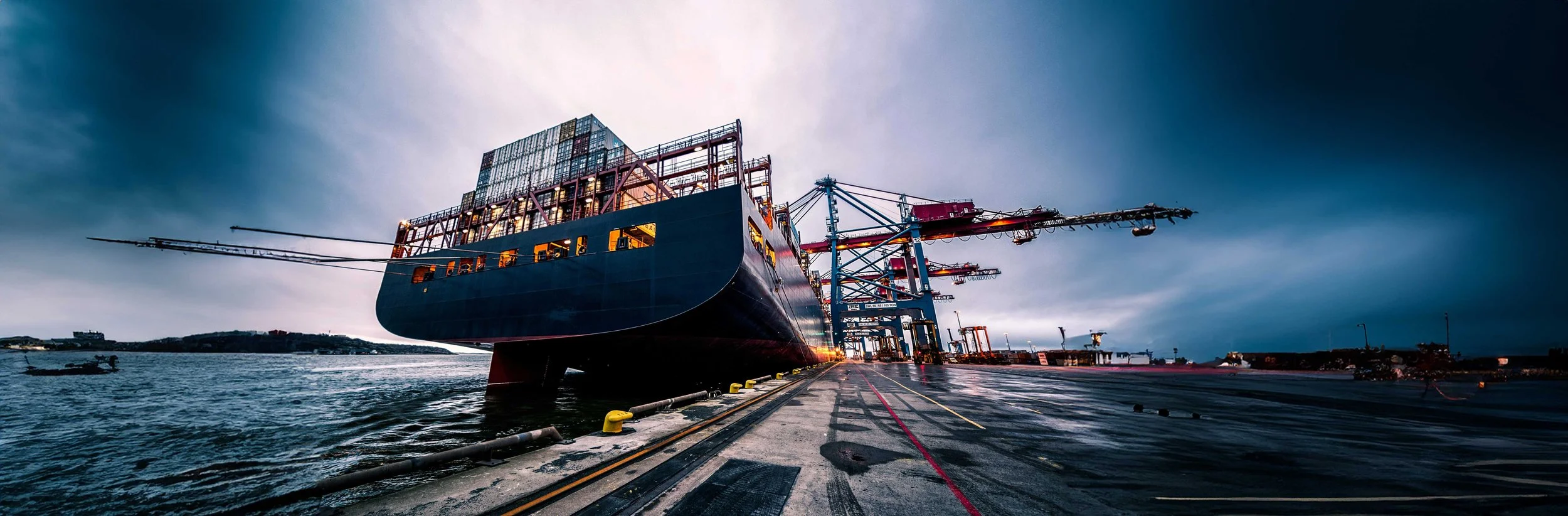 Large cargo ship docked at a port with cranes and shipping containers under a cloudy sky, illustrating The Weather Makers’ focus on resilient ports and maritime infrastructure through sustainable, nature-based engineering solutions.