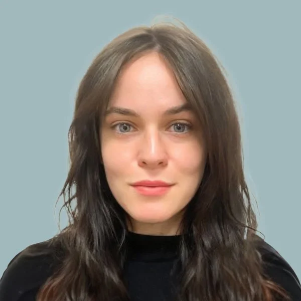 A young woman with long wavy brown hair and blue eyes, wearing a black top, standing against a light blue background.