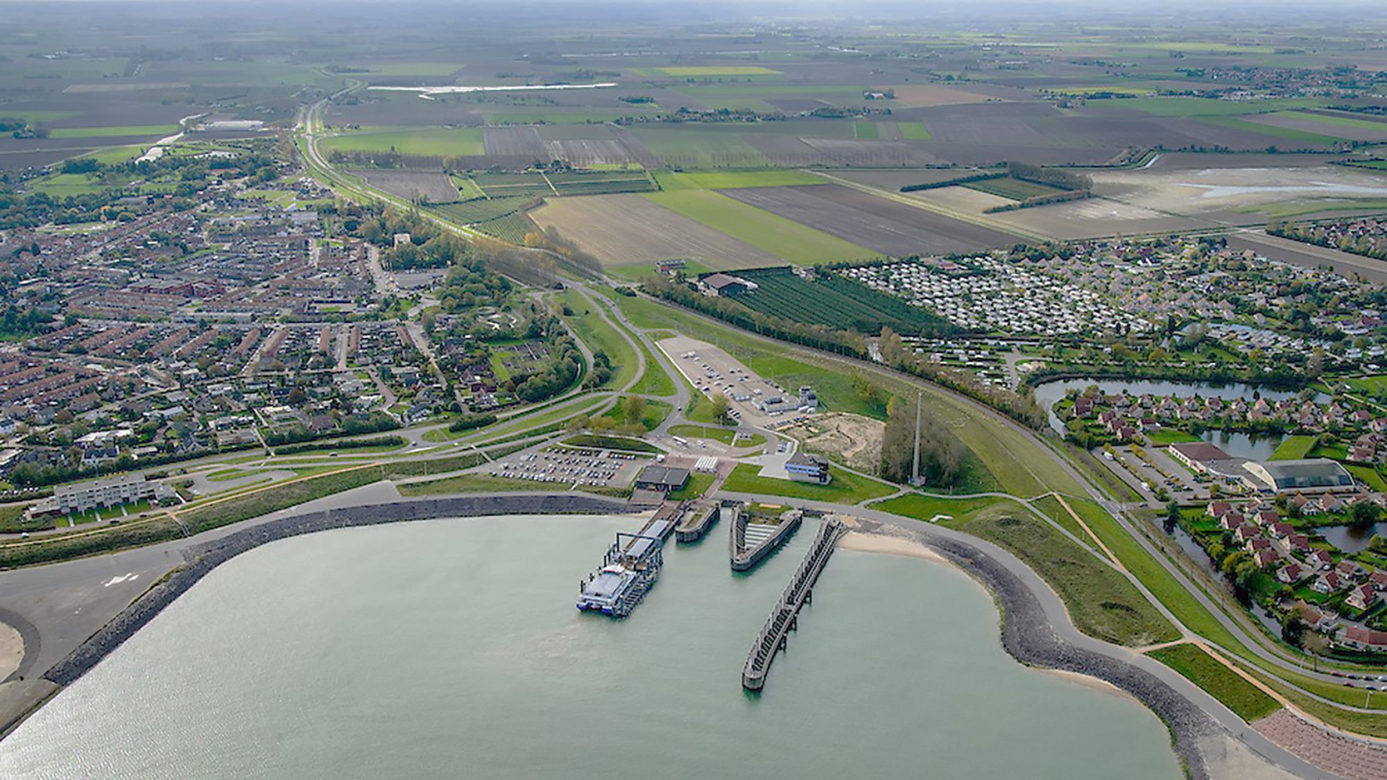 Aerial view of a dam in a town with boats docked at the marina, surrounded by residential neighborhoods, farmland, and green fields.