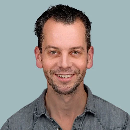Portrait of a smiling man with short dark hair, light beard, wearing a gray collared shirt against a light blue background.