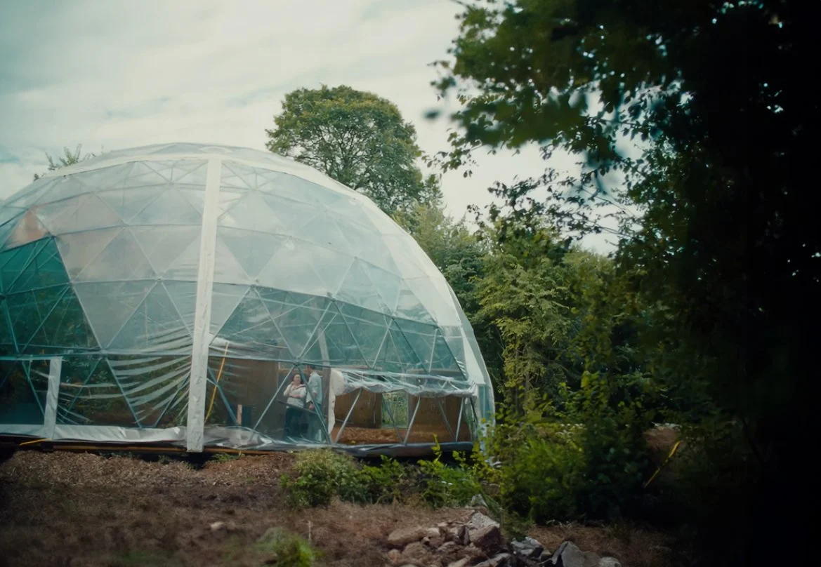 A transparent geodesic dome greenhouse in a wooded outdoor setting with trees and plants surrounding it.