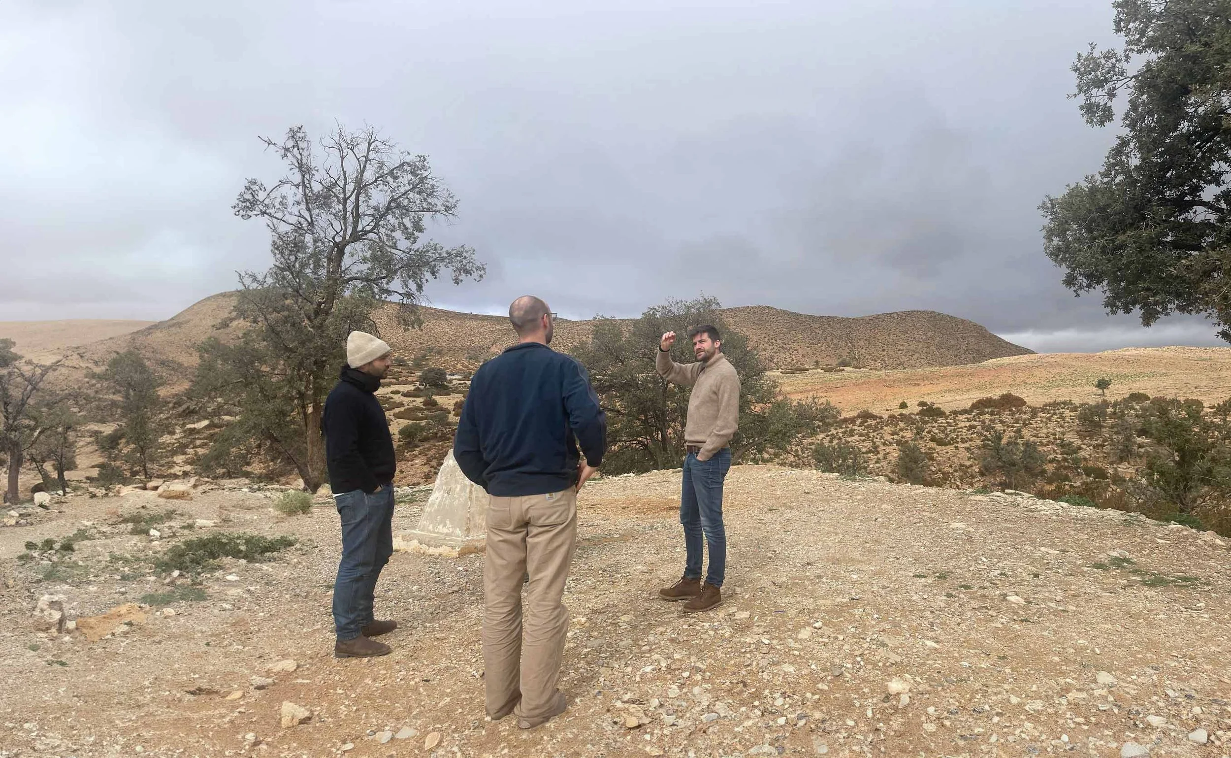 Three men standing outdoors in a rocky terrain with sparse trees and hills in the background, under a cloudy sky, engaging in conversation.