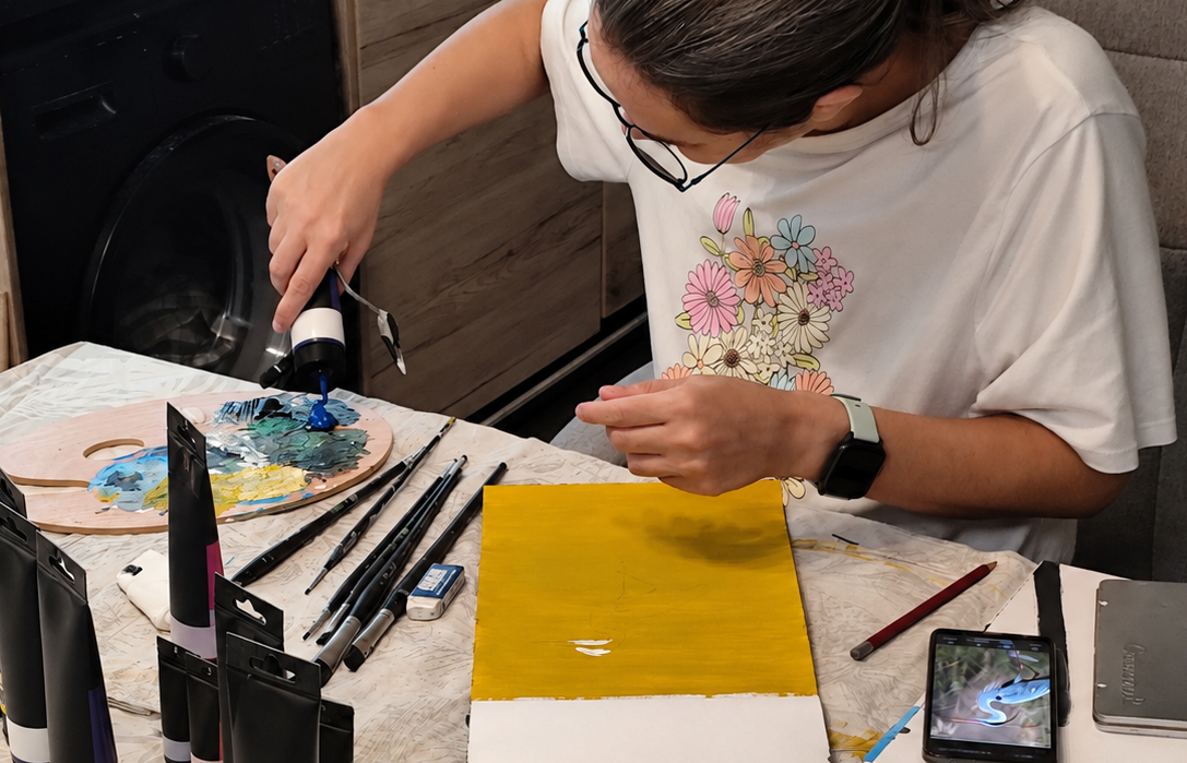 Chaya creating artwork at a table with brushes, paints, and a yellow canvas.