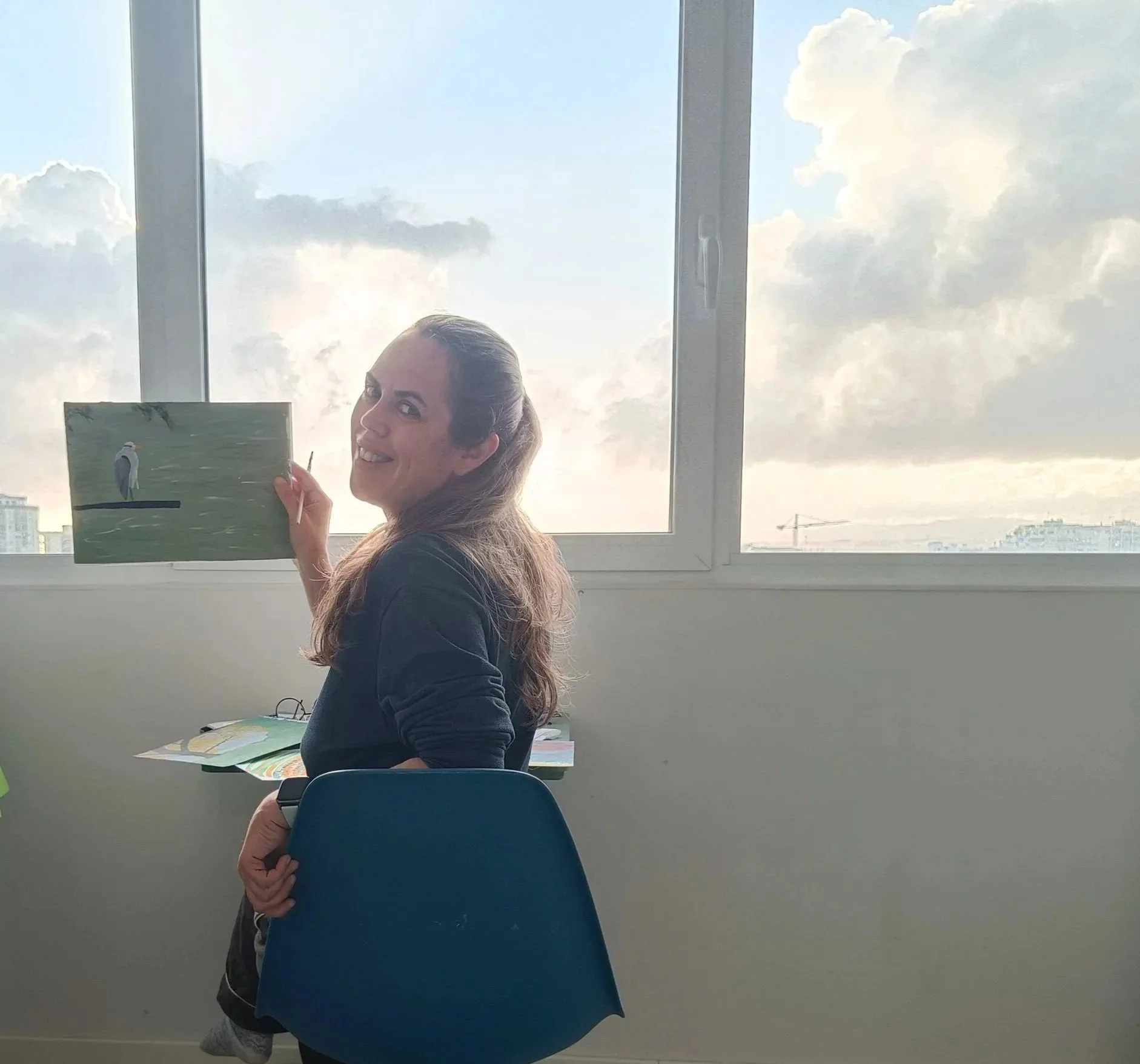 Chaya holding her acrylic painting “Heron in Stillness” in front of a window overlooking the city.