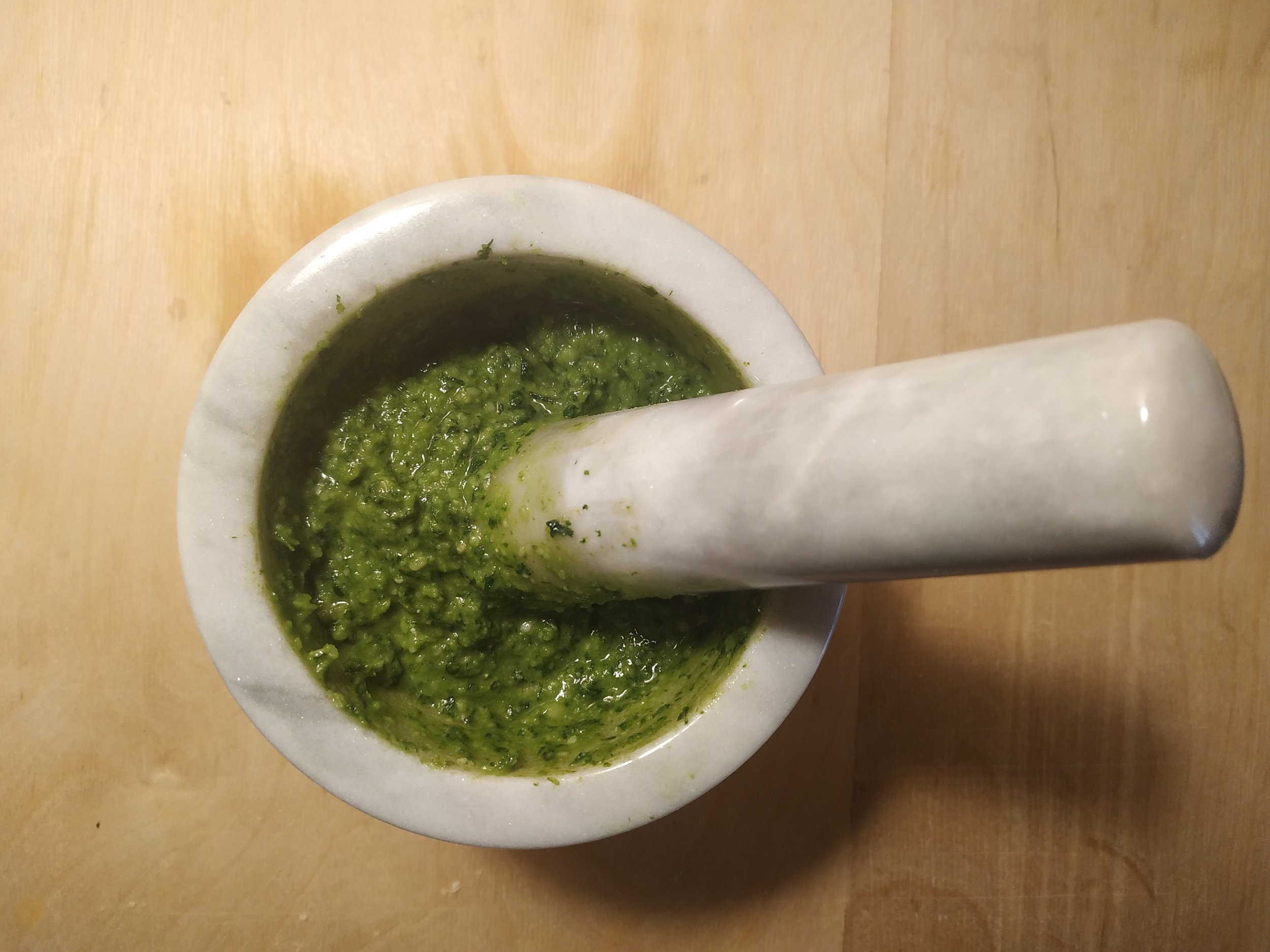 Fresh pesto being prepared with a mortar and pestle on a wooden surface.
