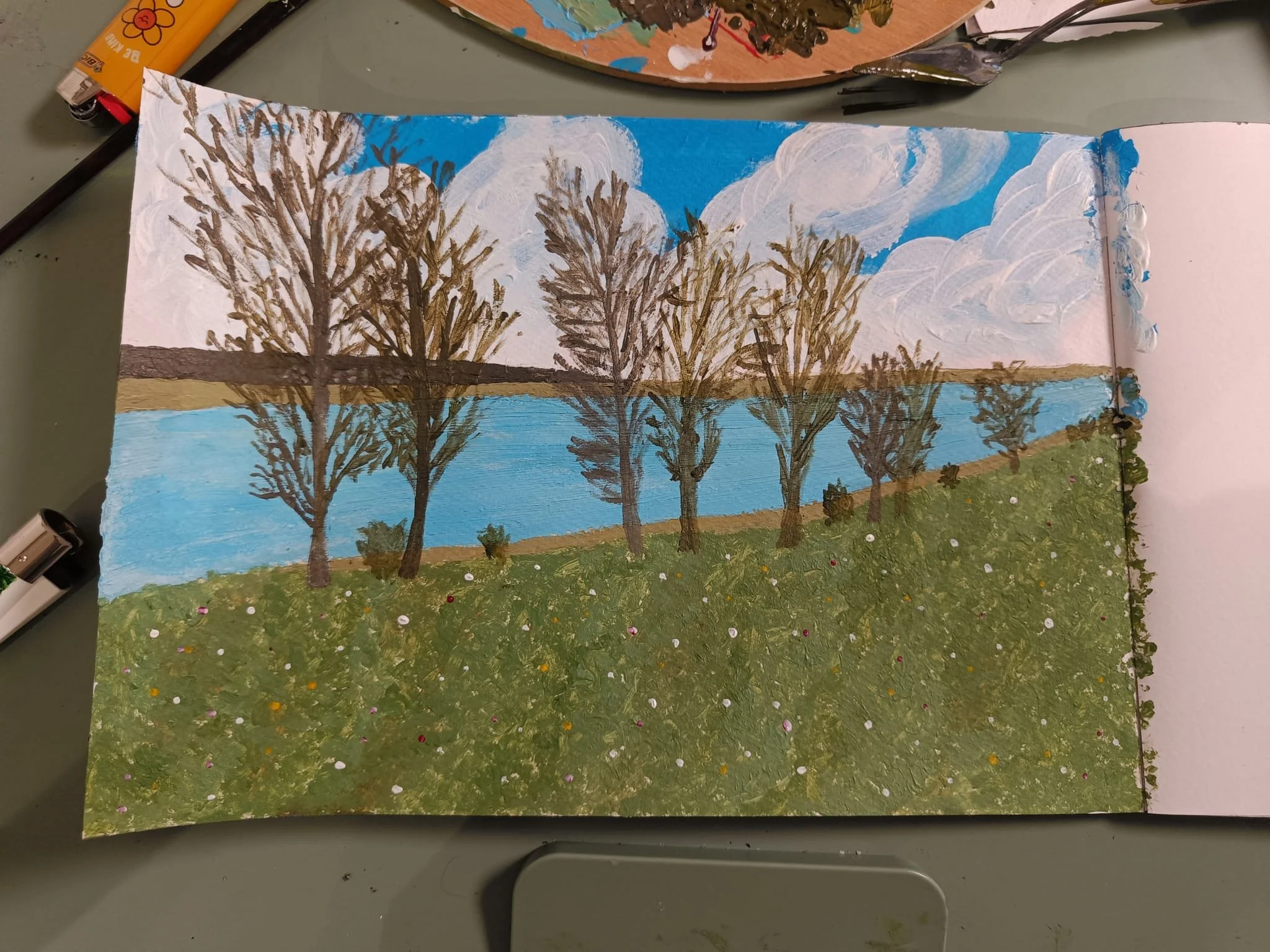“Biking by the Lake,” an acrylic painting by Chaya showing leafless trees beside a lake, green grass, and a partly cloudy sky.