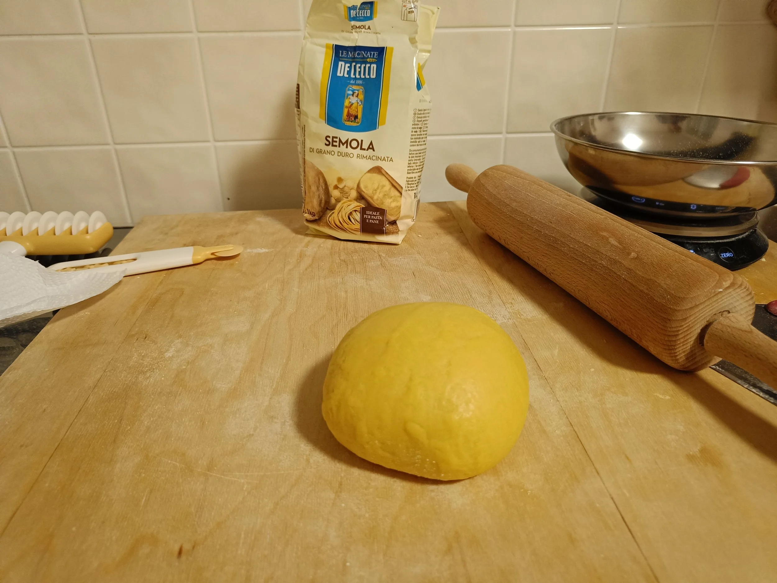A ball of fresh pasta dough on a wooden board, with semolina flour, a rolling pin, and kitchen tools nearby.