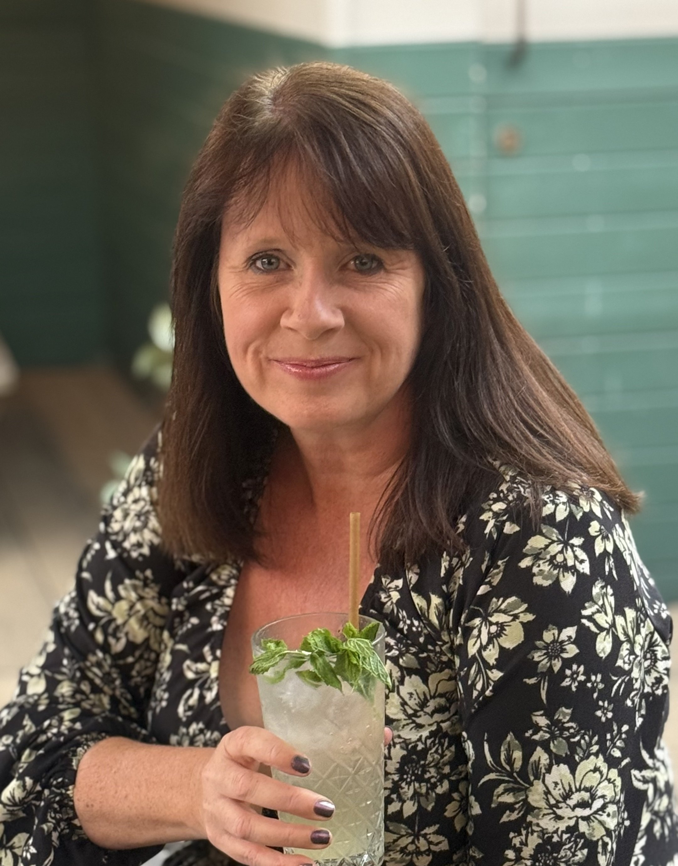 A woman with brown hair and wearing a black and white floral patterned dress is holding a mint mojito cocktail with a straw and fresh mint leaves.