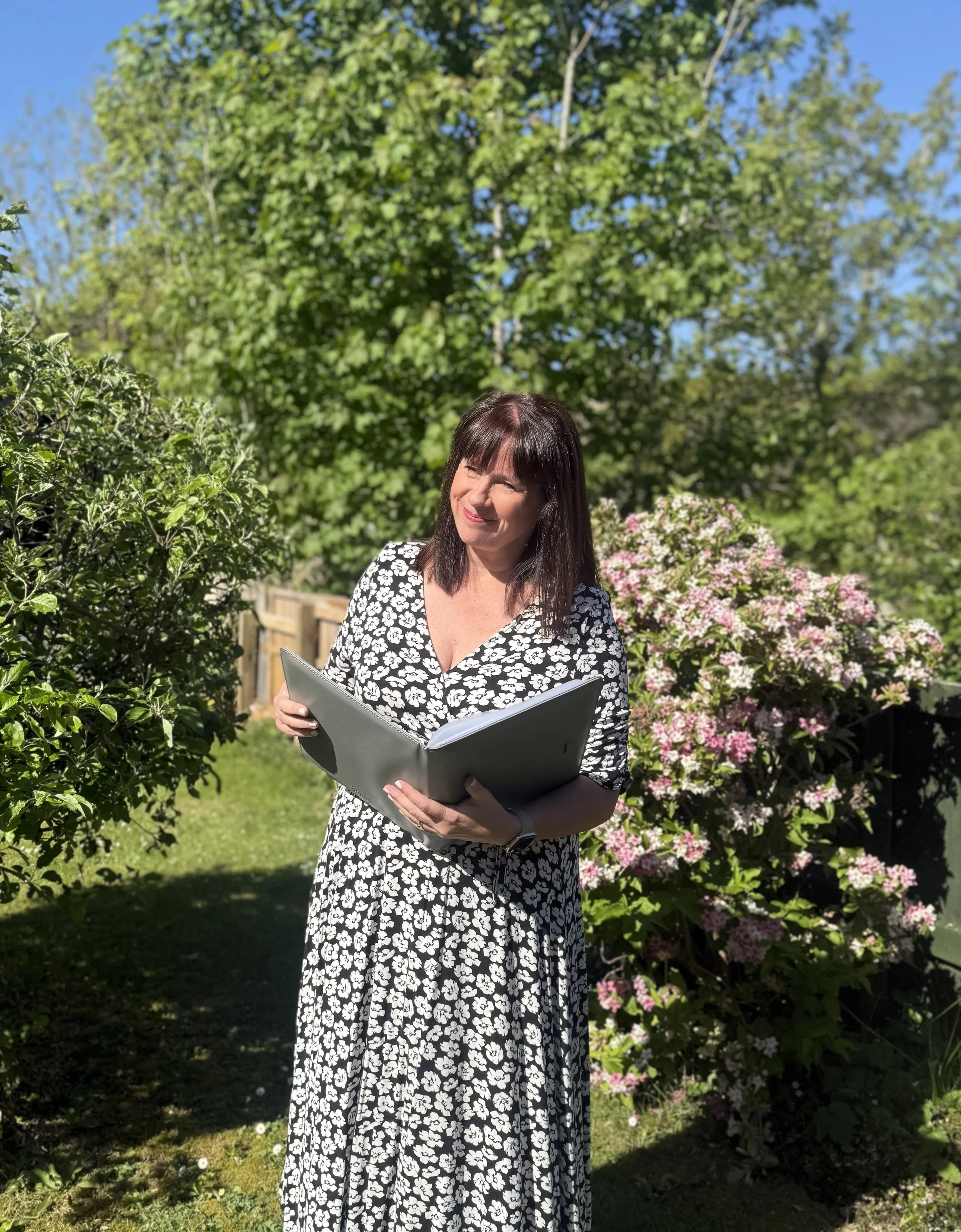A woman in a black and white floral dress standing outdoors in a garden, holding a folder or book, with various green trees and pink flowering bushes in the background on a sunny day.
