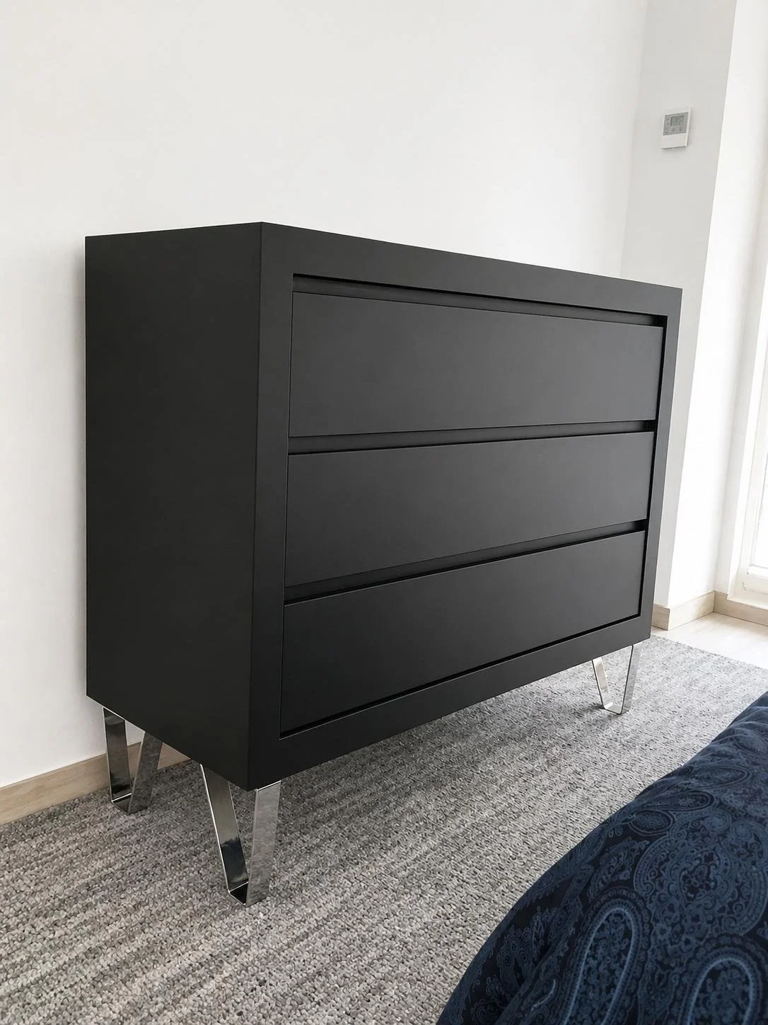 Commode noire moderne avec trois tiroirs, pieds en métal argenté, dans une pièce avec mur blanc et tapis gris.