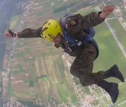 A person skydiving with a yellow helmet, open arms, and a joyful expression, free-falling above farmland and rural scenery.