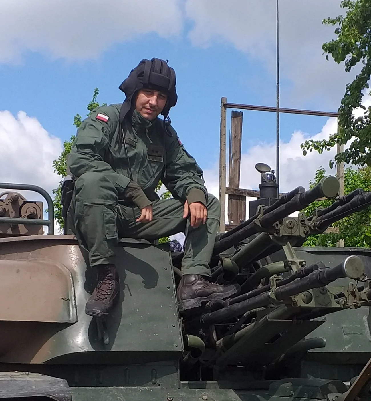 A soldier in military uniform sitting on a military vehicle with mounted guns, wearing a helmet and tactical gear, outdoors under a partly cloudy sky.