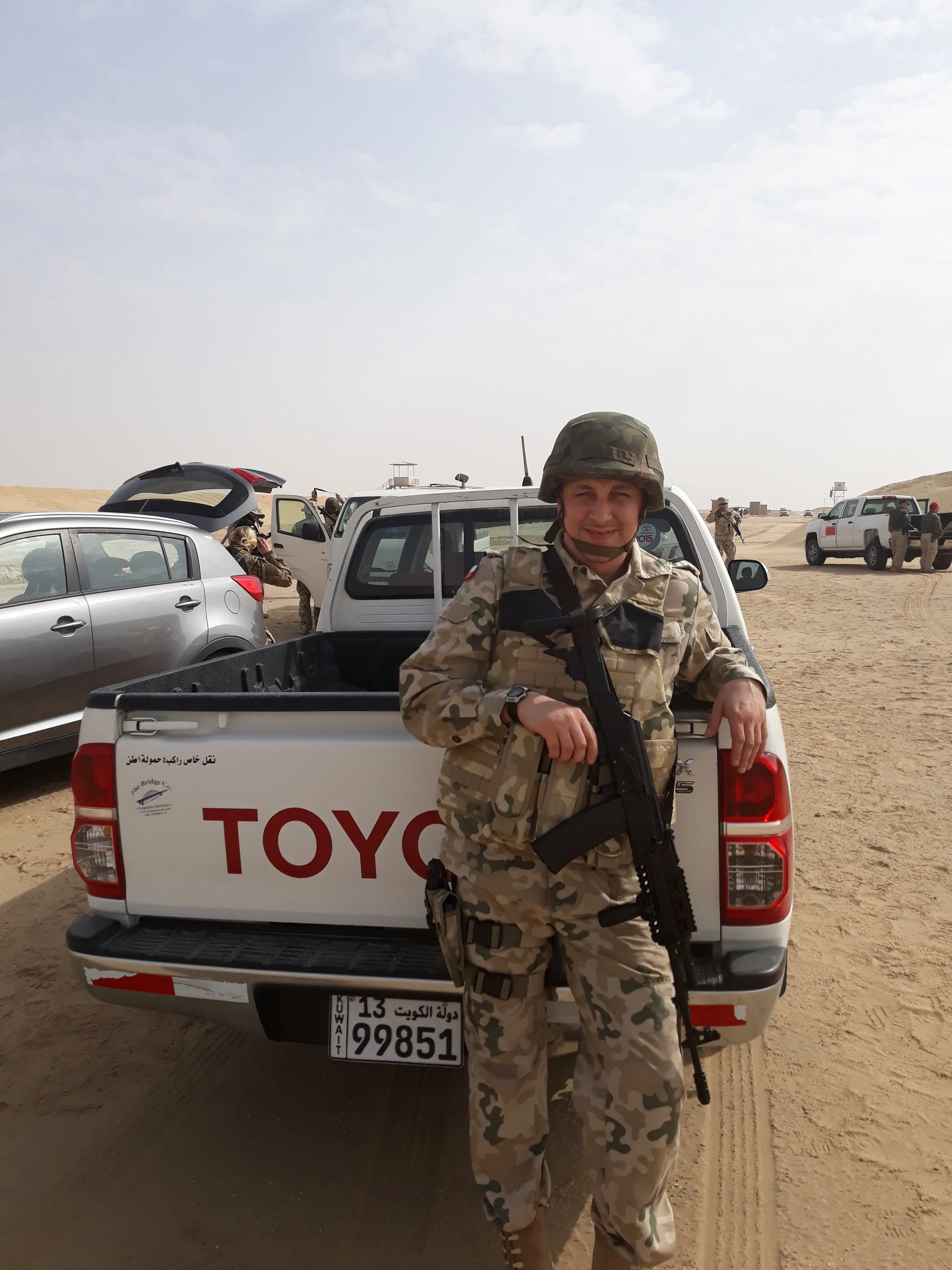 A soldier in camouflage uniform holding a rifle standing in front of a white pickup truck with Kuwaiti license plates in a desert landscape with other military personnel and vehicles in the background.