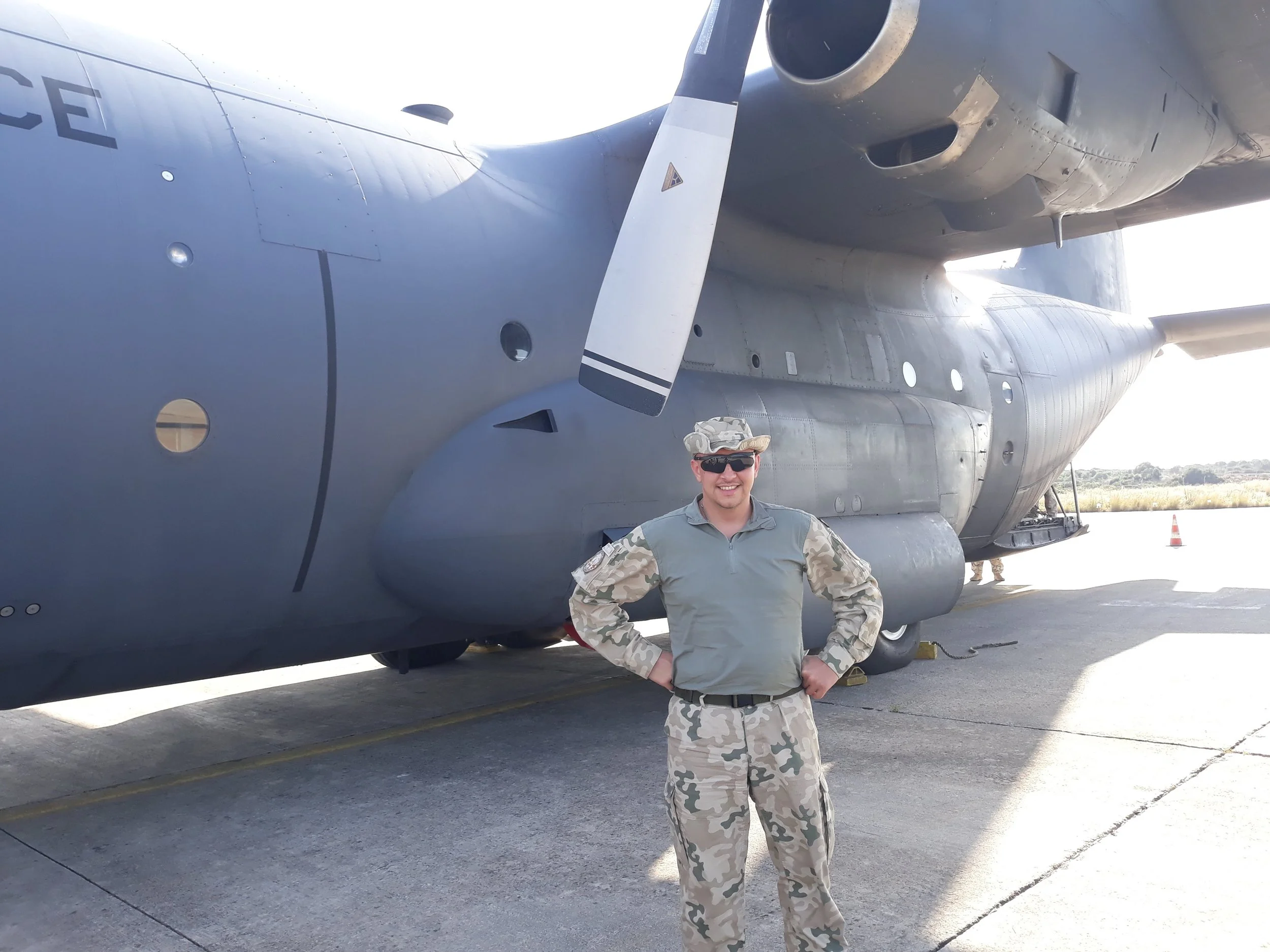 A man in military camouflage uniform and sunglasses standing in front of a large military aircraft, smiling.