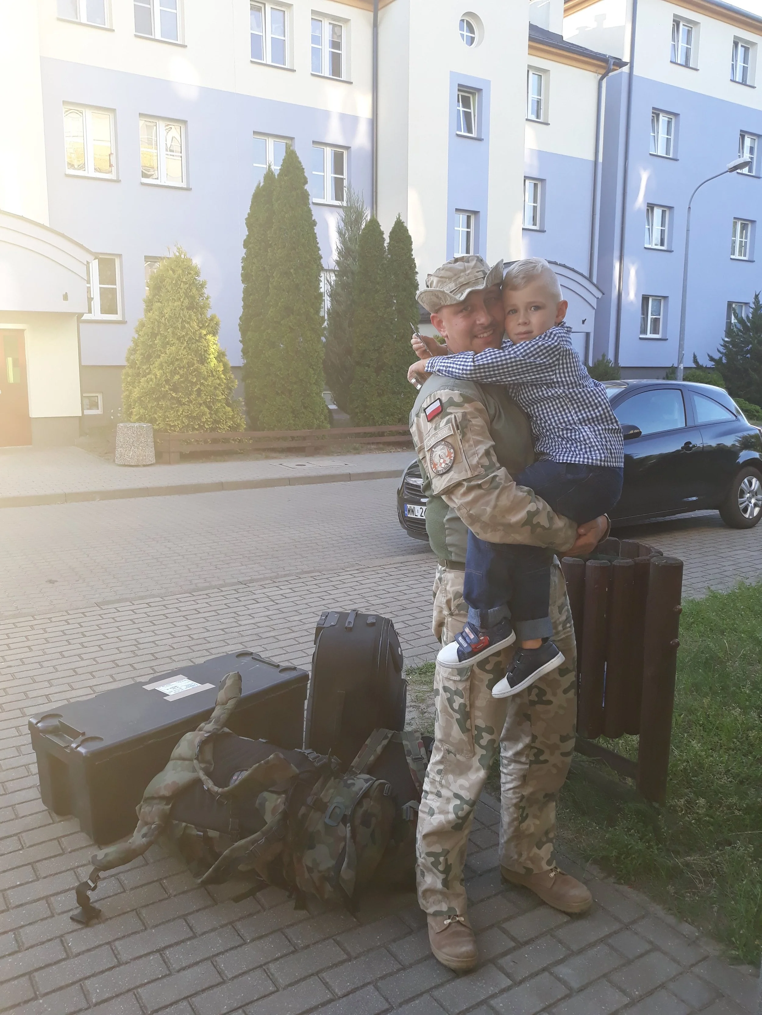 A soldier in camouflage uniform holding a young boy in front of a residential building with trees. There are two black suitcases and a tactical bag on the ground.
