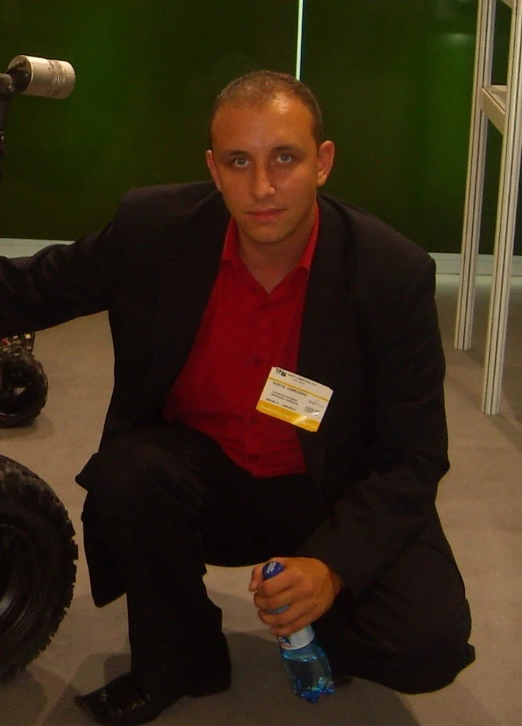 A man with short hair, wearing a black suit and a red shirt, squatting indoors next to a green wall. He is holding a blue water bottle and has a name badge clipped to his suit. There are some equipment and a partial view of a vehicle tire near him.