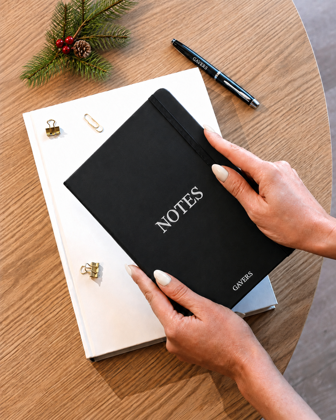 A person's hands holding a black notebook labeled 'NOTES' on a wooden desk. On the desk, there are white paper clips, gold binder clips, a black pen, a white notebook, and a small sprig of pine with red berries and pine cone.