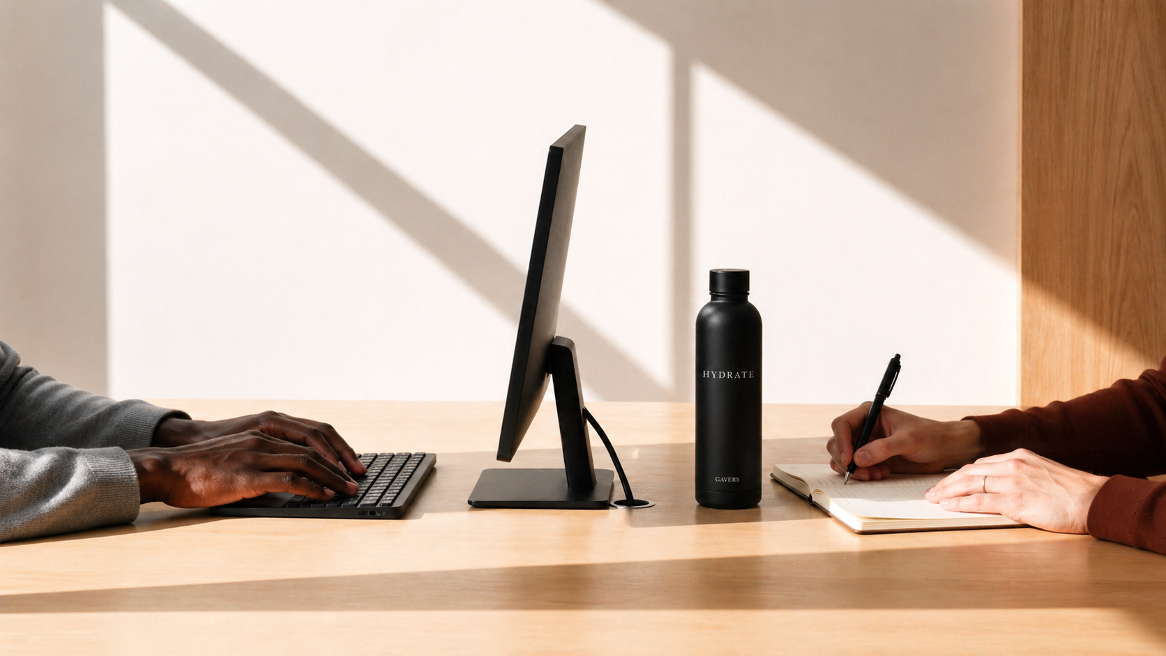 Person on the left typing on a keyboard, and person on the right writing in a notebook, with a black water bottle labeled 'HYDRATE' and a computer monitor in between on a wooden desk.