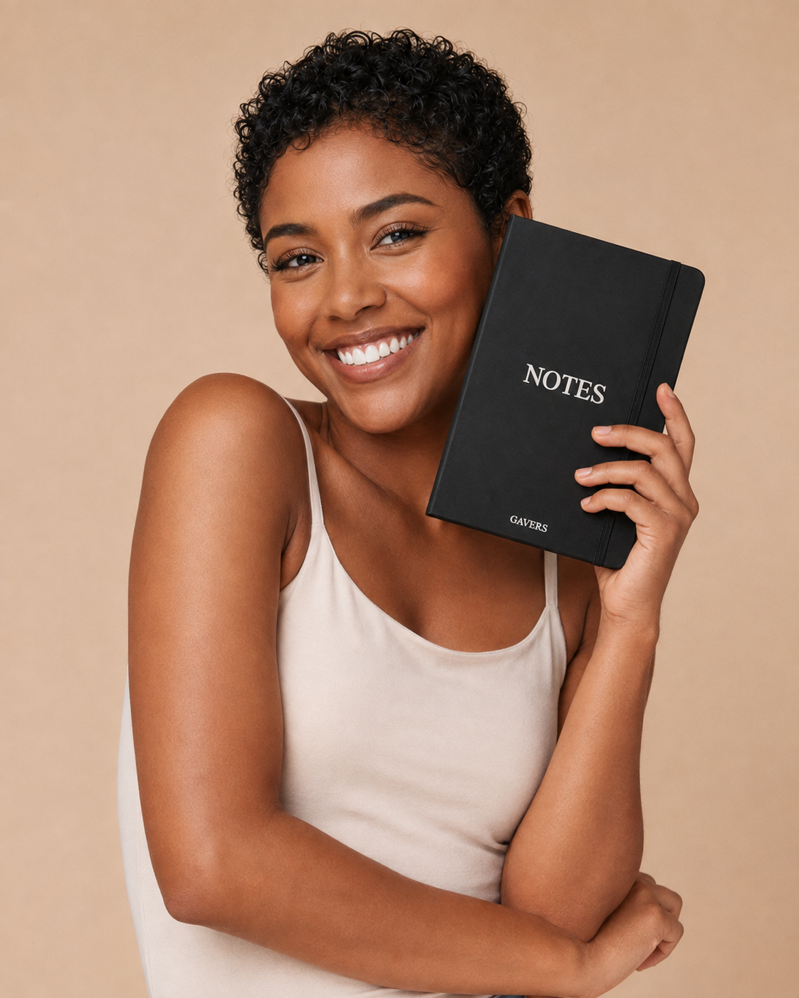 Smiling woman with short curly hair holding a black notebook labeled notes close to her face, wearing a beige sleeveless top, against a beige background.