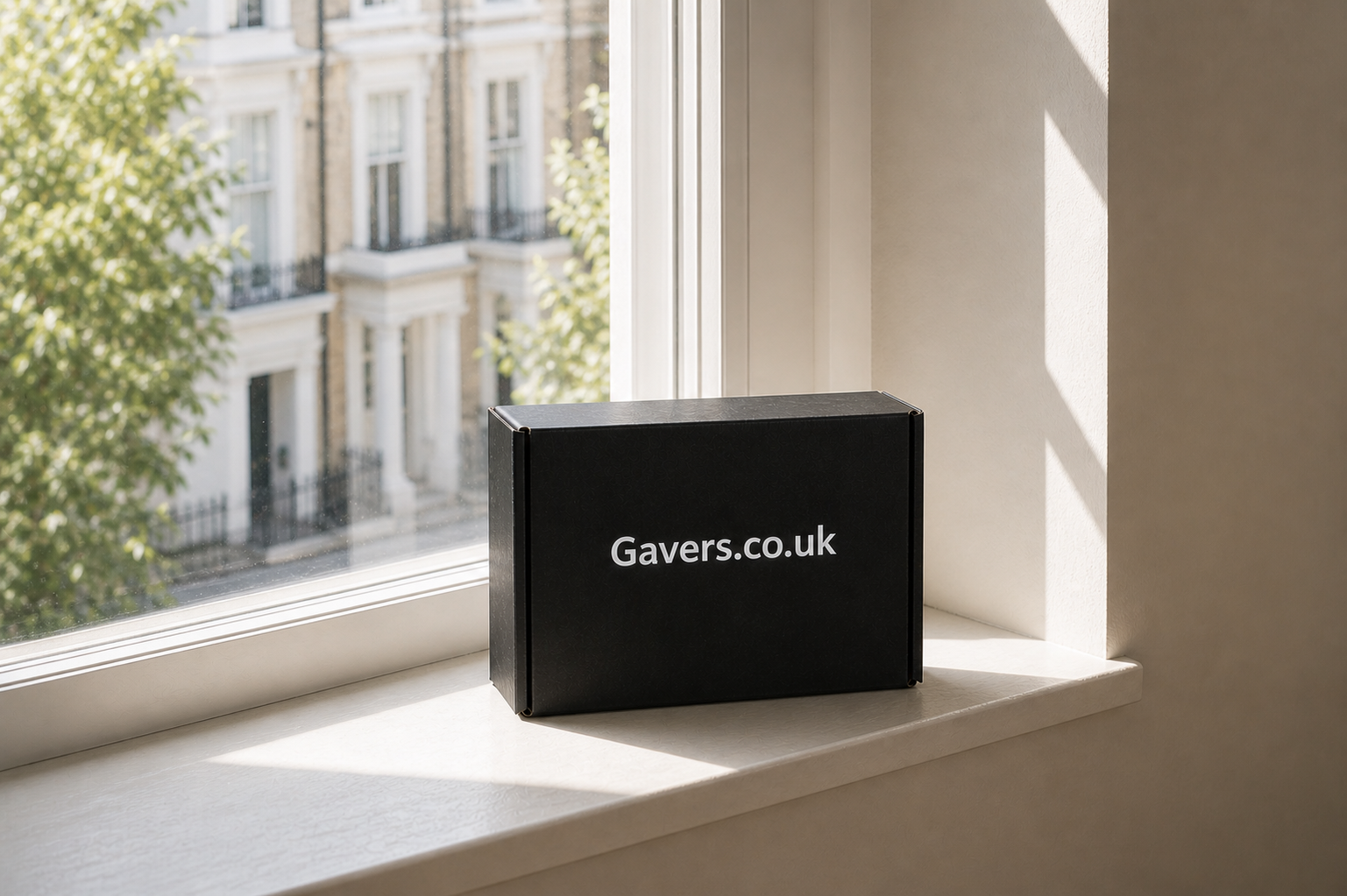 A black box with the text "Gavers.co.uk" on it, placed on a windowsill with a view of a building with balconies outside.