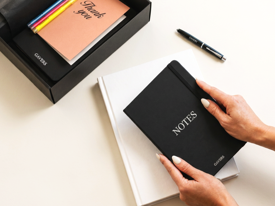 A person holding a black notebook labeled 'NOTES' above a white notebook, on a white desk, with a black pen nearby and a black gift box containing colorful pens and a pink 'Thank you' card.