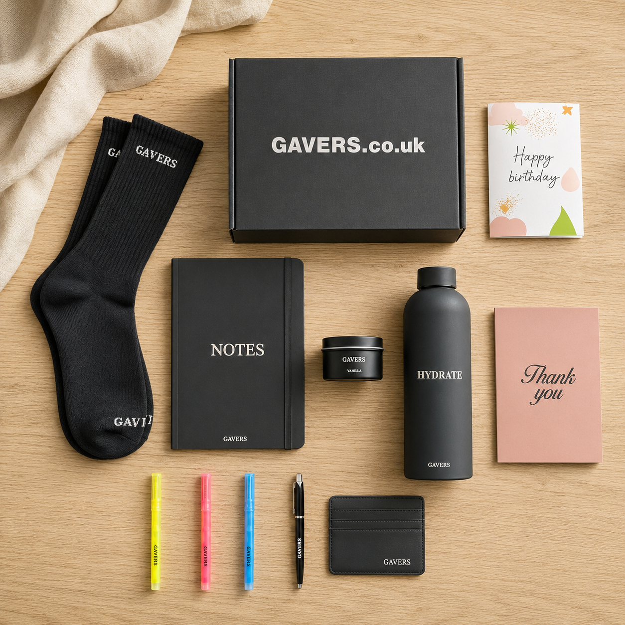 Gavers.co.uk gift box along with various branded items including black socks, notebook, notes, water bottle, small tin of Gavers Vanilla product, hydration bottle, thank you card, birthday card, colorful pens, a pen, a cardholder, and a compliment card on a wooden surface with a beige fabric in the corner.