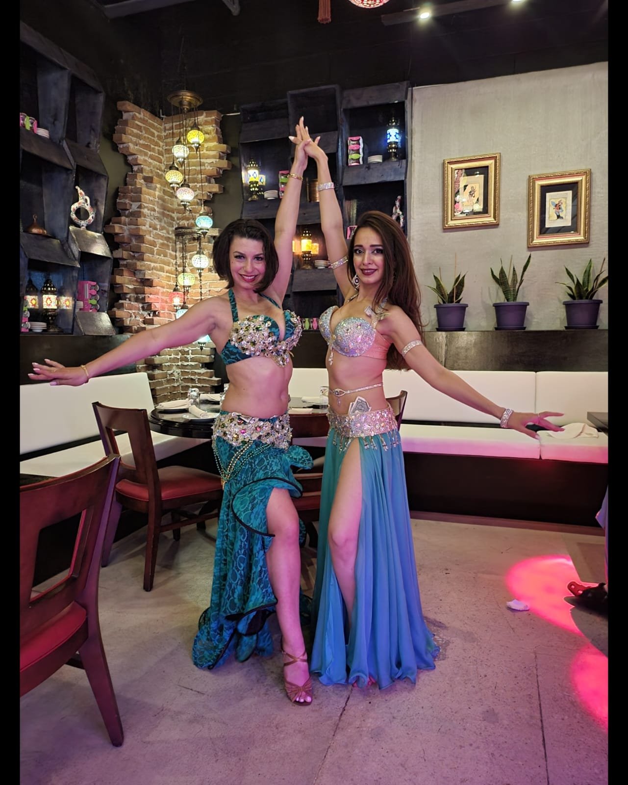 Two women in colorful, embellished costumes dance inside a restaurant. One woman is standing on a chair and the other is standing on the floor, holding their hands high. The restaurant has a brick wall with hanging lanterns, framed pictures, and pott