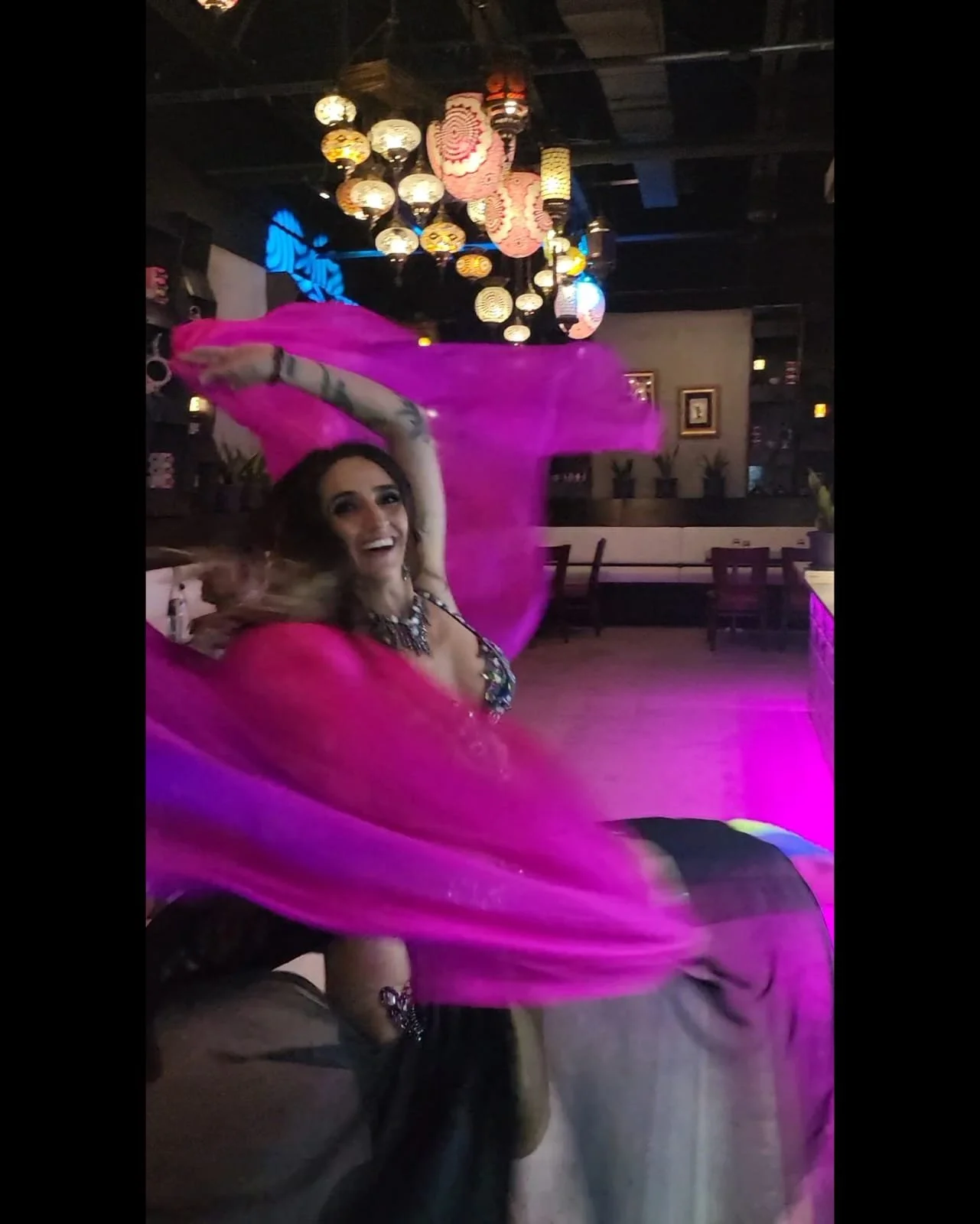 A woman smiling and dancing with pink fabric swirling around her inside a dimly lit venue decorated with colorful hanging lanterns.