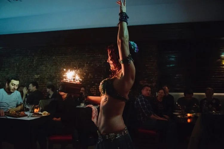 A belly dancer holding a cake with sparklers in a dimly lit restaurant with seated patrons.